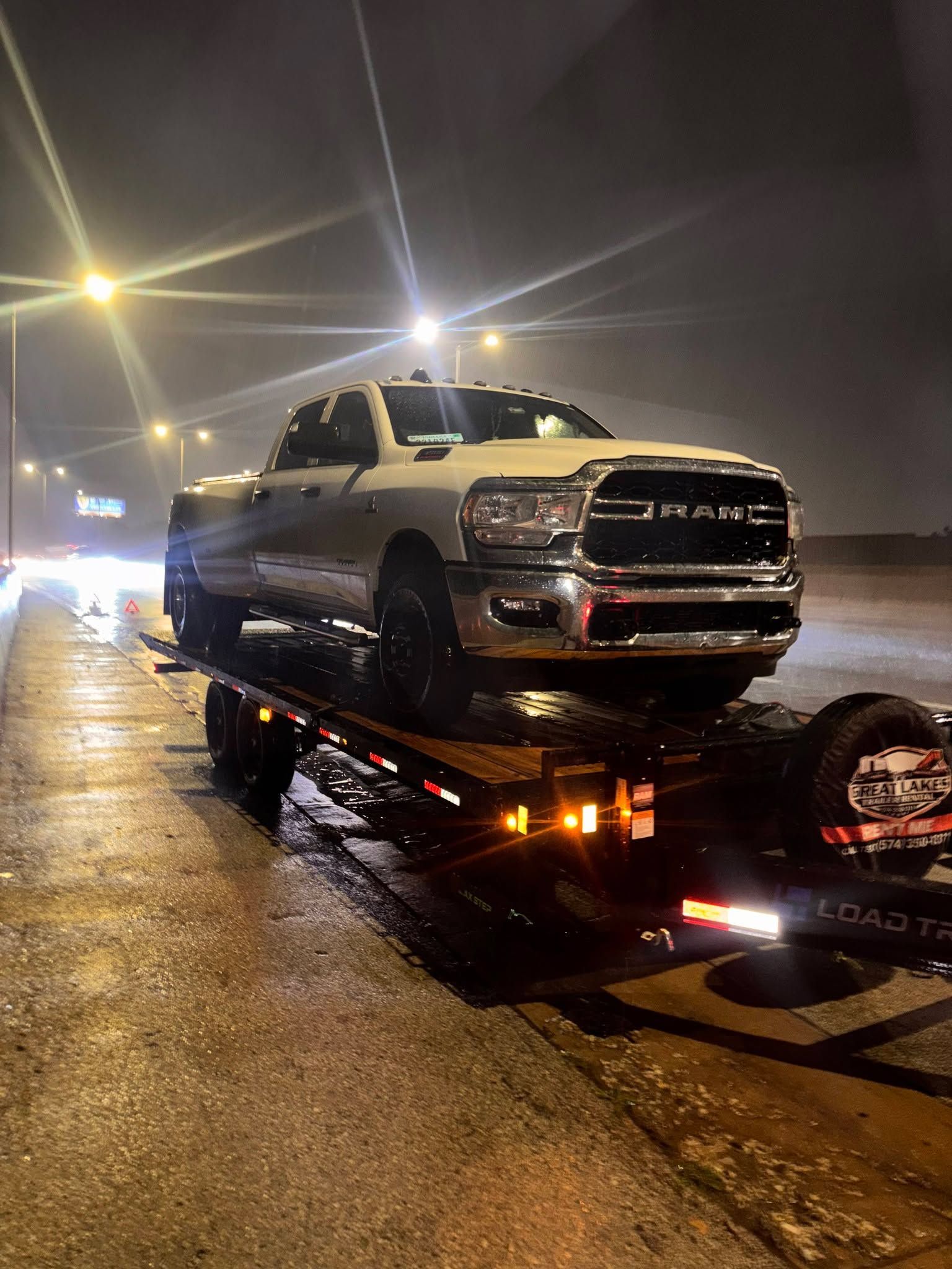 Silver pickup truck on a flatbed tow trailer at night on a wet, rainy street