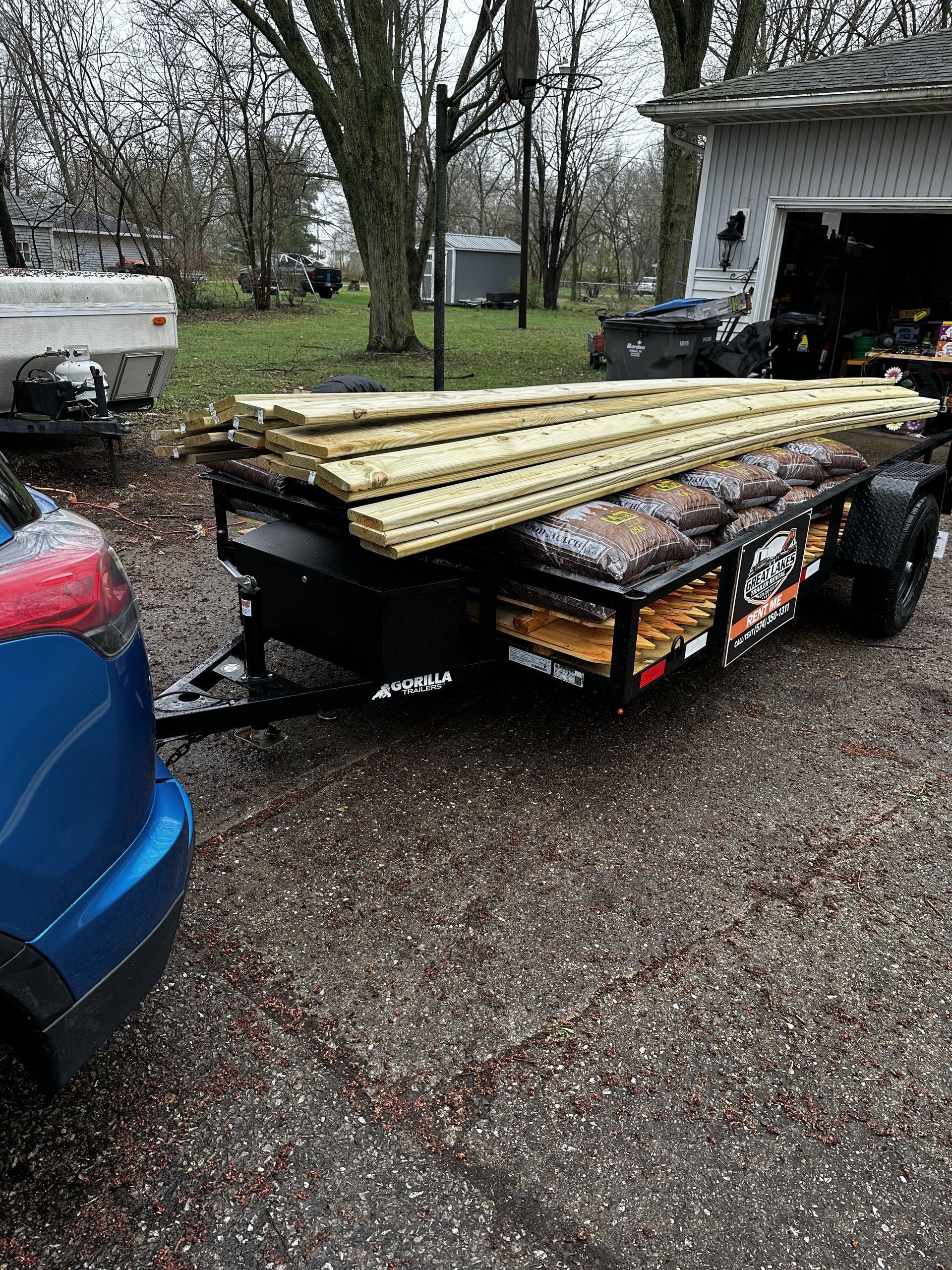 Black utility trailer loaded with lumber in a wet driveway beside a blue car and garage.