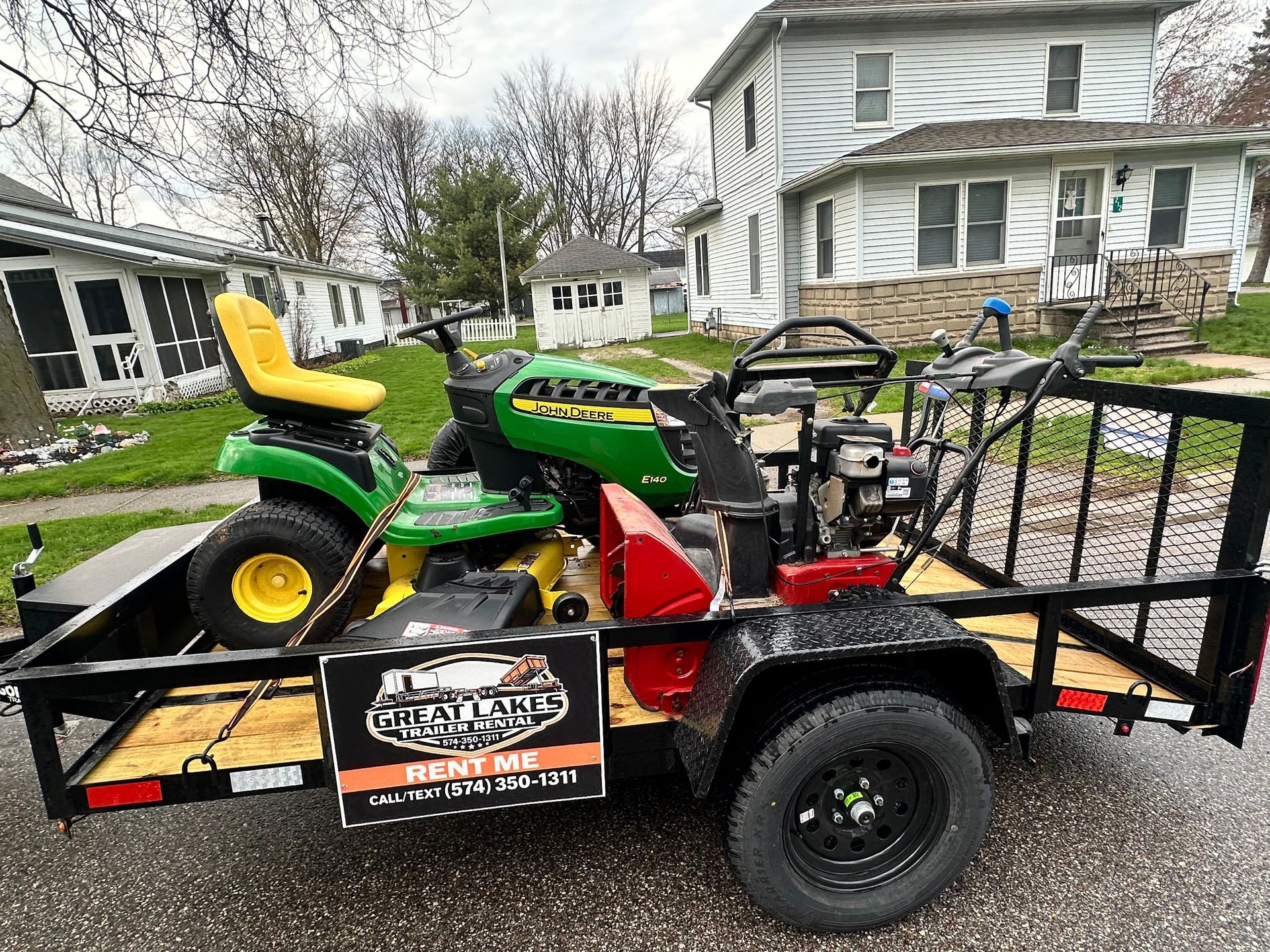 Green John Deere lawn tractor and trailer loaded on a black utility trailer in a residential yard.
