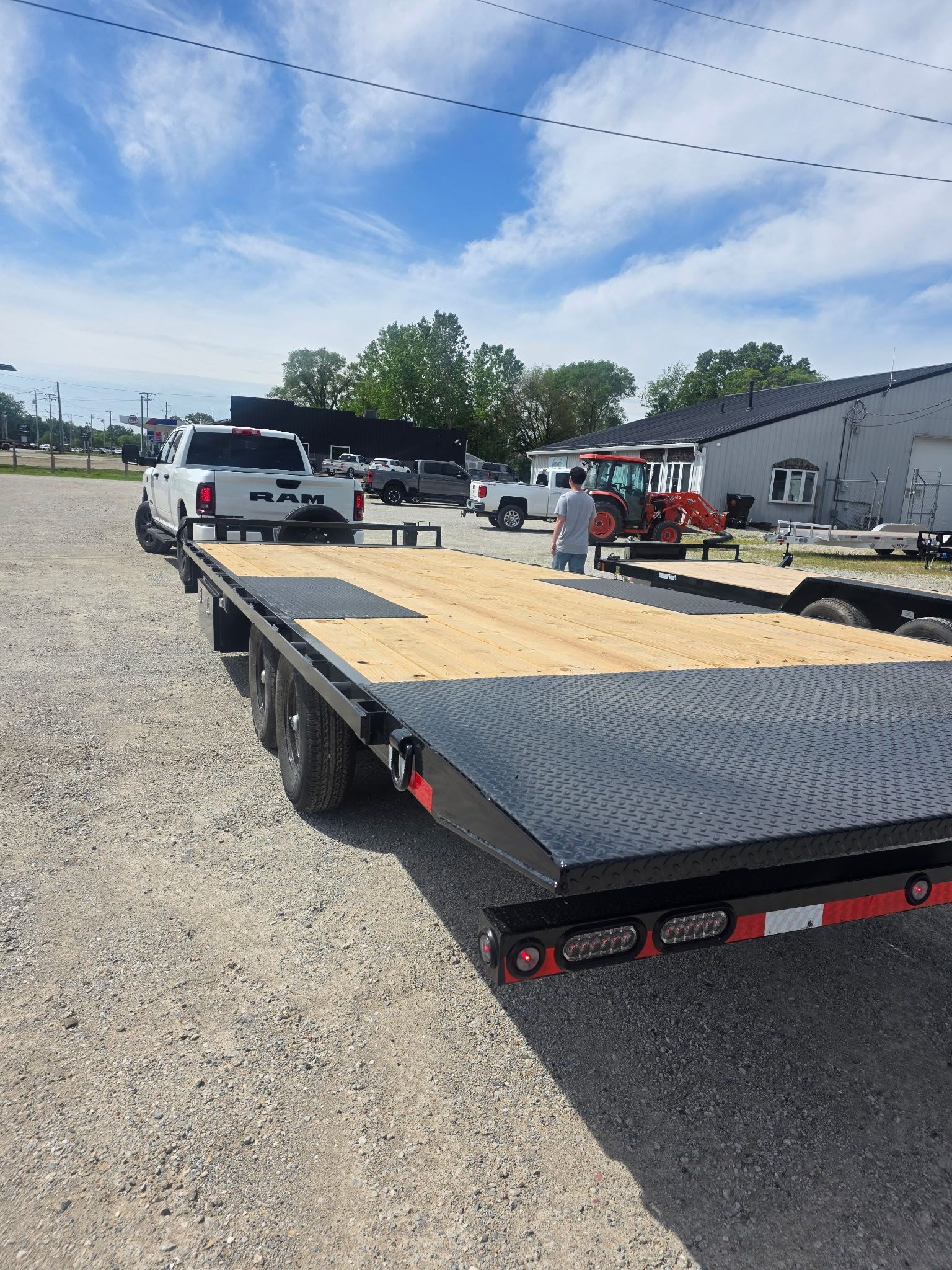 Flatbed trailer in gravel lot with white pickup trucks and a red forklift under a blue sky.