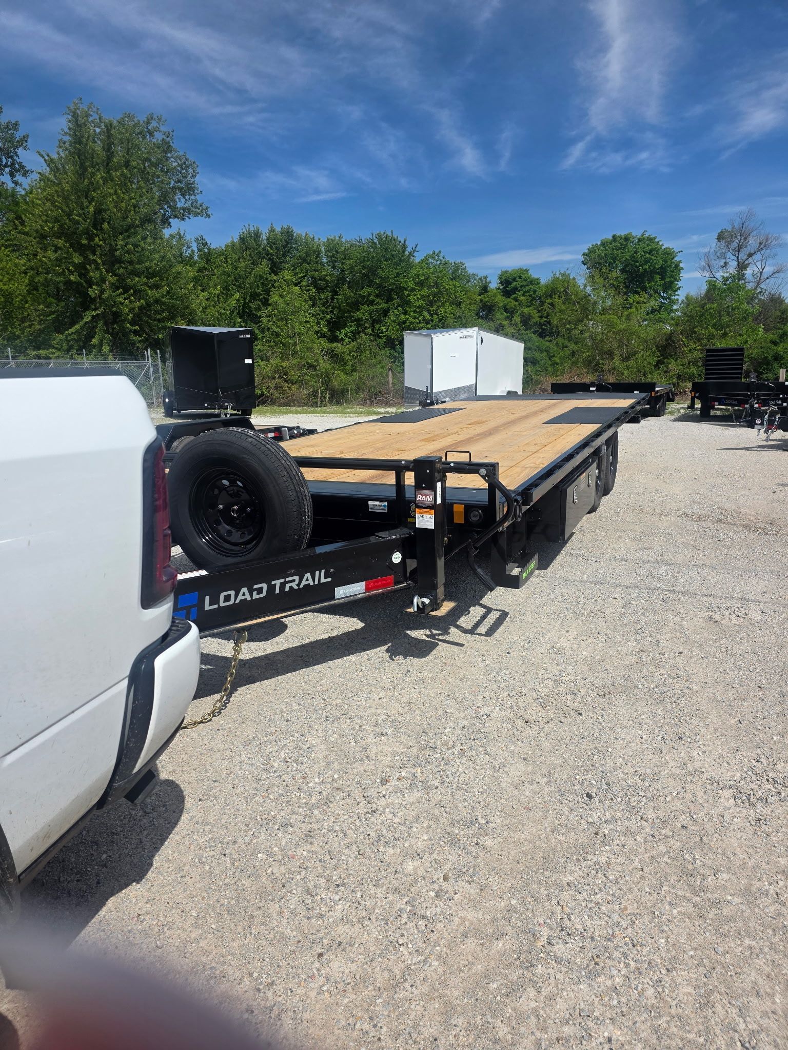 Flatbed trailer loaded with plywood beside a white truck on a gravel lot under a blue sky