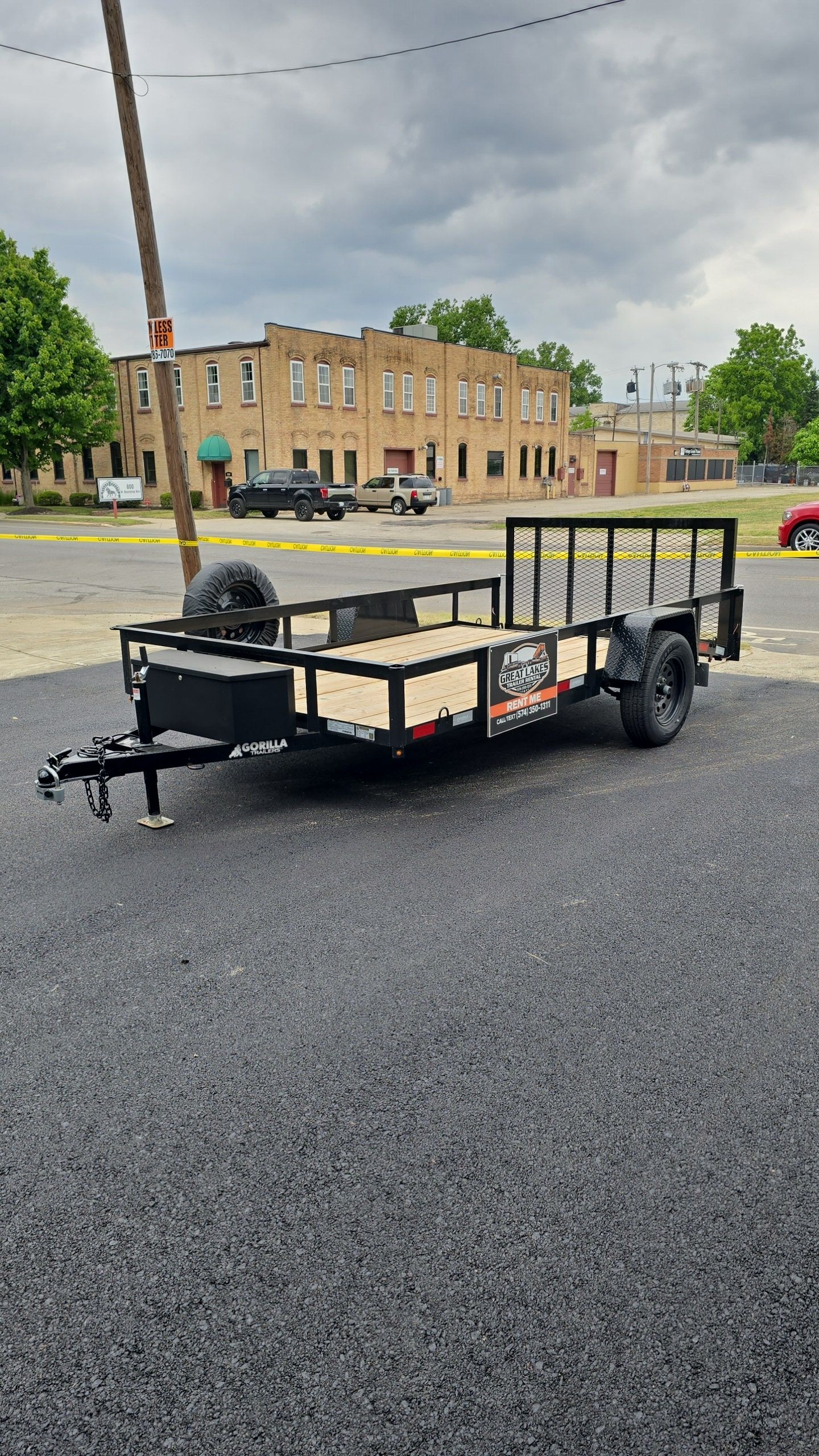 Flatbed utility trailer with black frame and tan sides parked on a gravel lot near brick buildings.