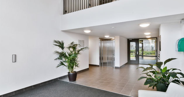 Bright apartment lobby with potted plants, tiled floor, white walls, and glass doors ahead