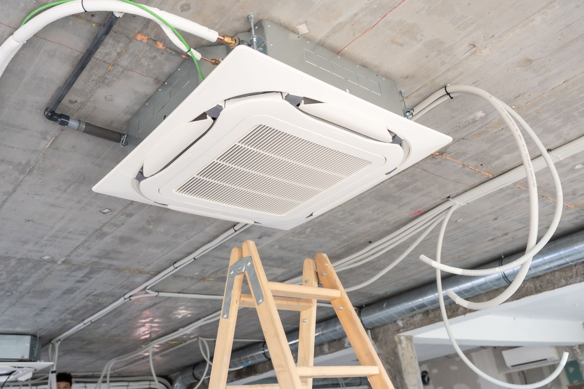 Ceiling-mounted HVAC unit installed in a concrete ceiling during construction, with a ladder nearby and exposed wiring.