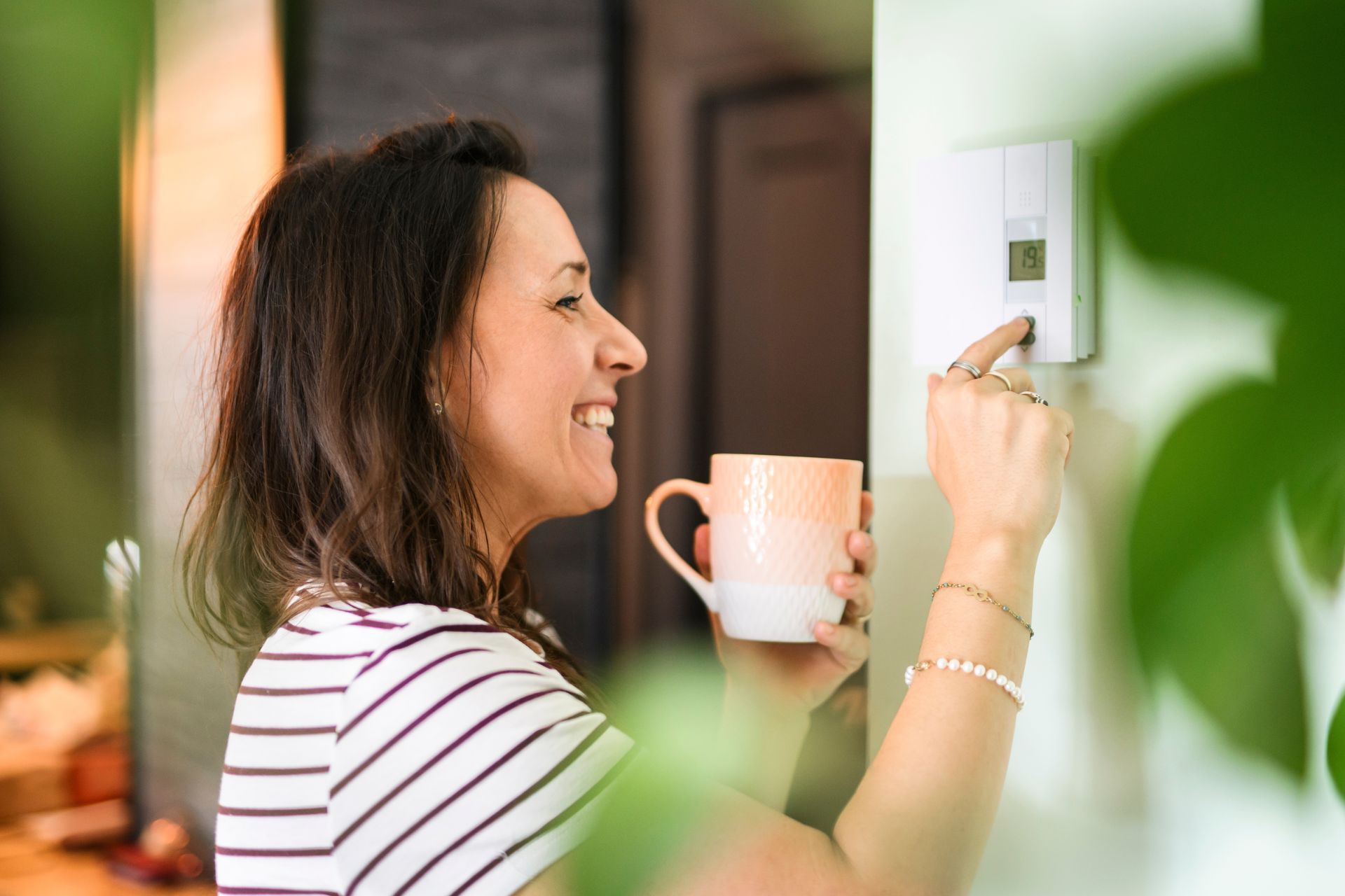 Woman smiling while adjusting a wall thermostat and holding a mug in a bright home interior