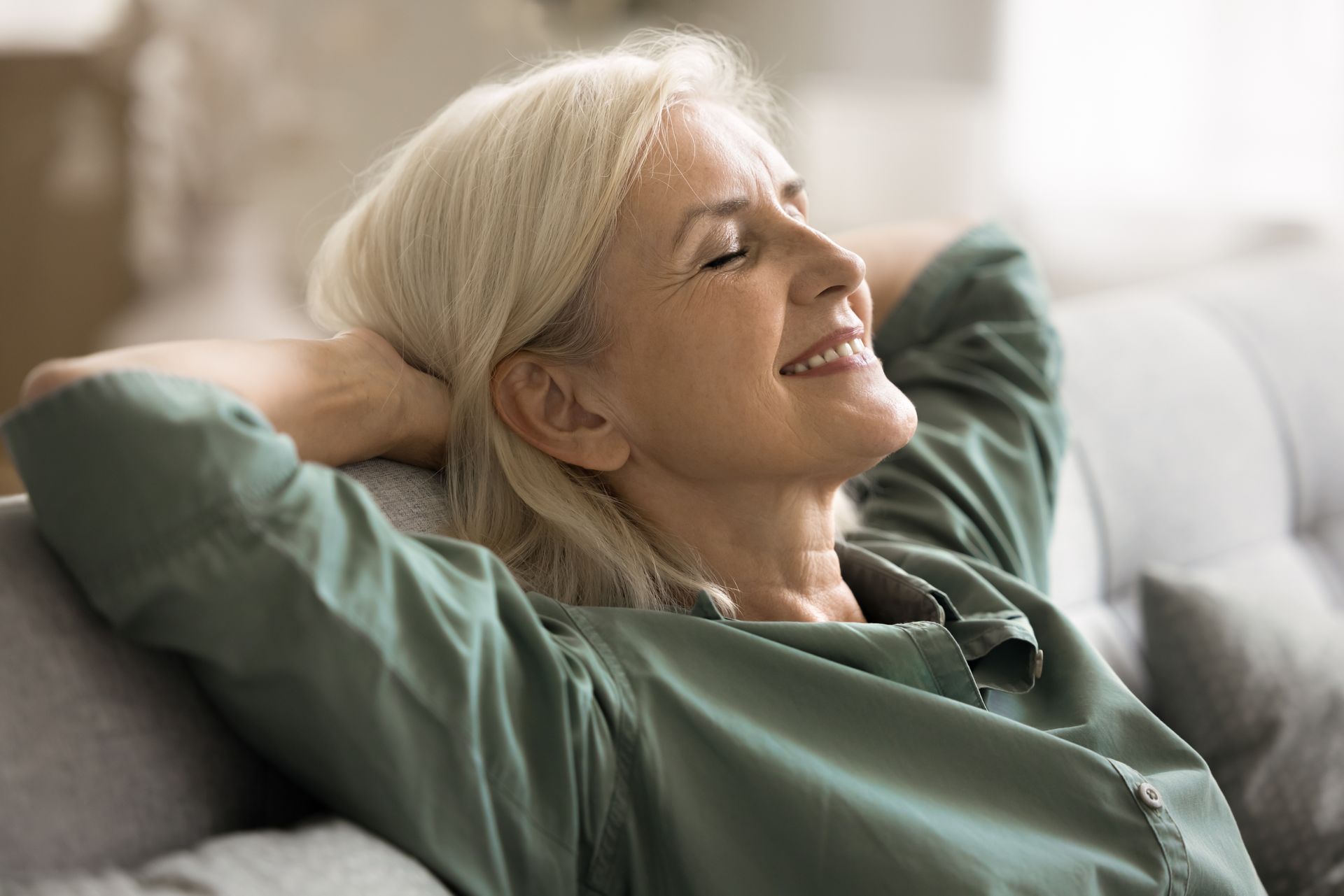 Relaxing woman reclines on a sofa, smiling with hands behind her head, wearing a green blouse.
