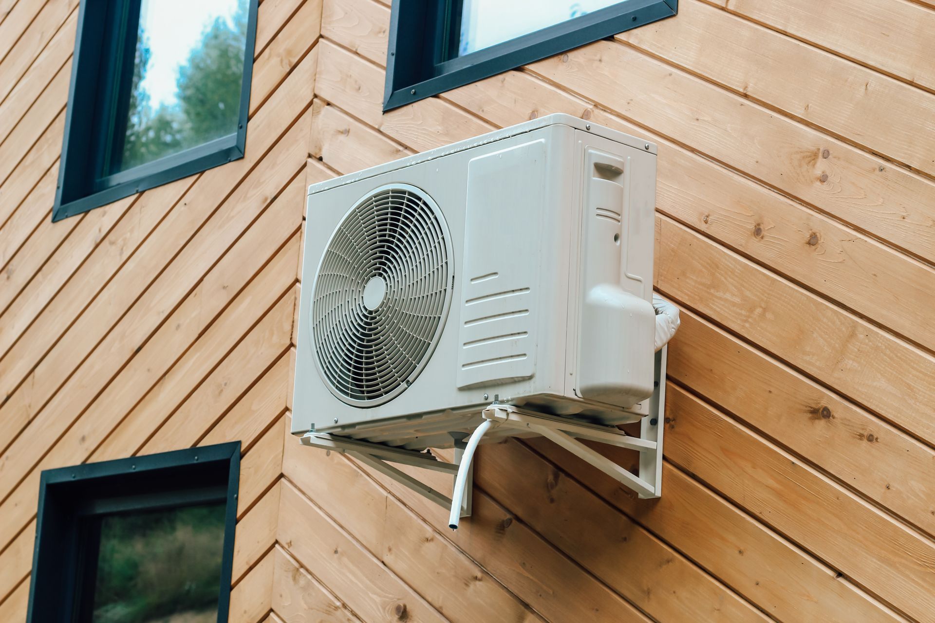 Outdoor air conditioner unit mounted on a wooden house wall beneath windows