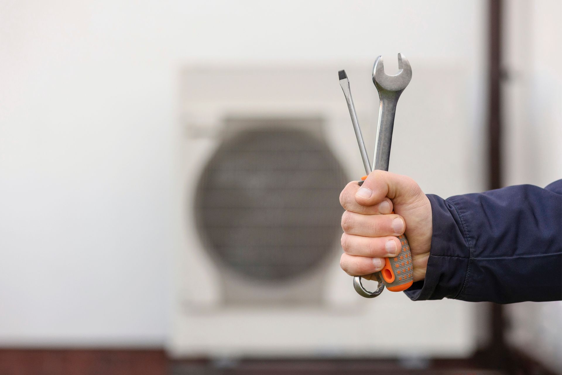 Hand holding a wrench and screwdriver in front of an outdoor air-conditioning unit
