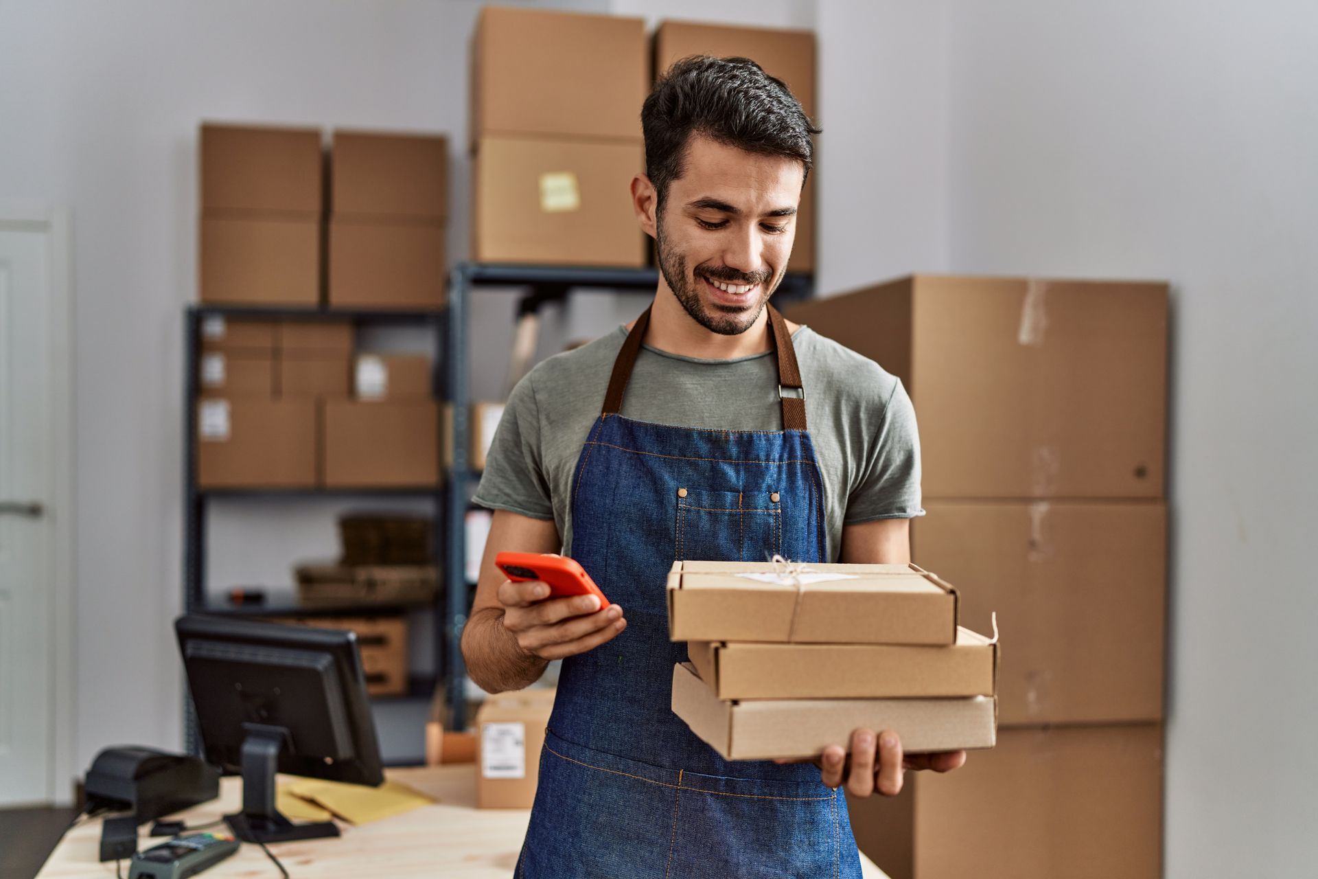 Man in apron smiles, looking at phone, holding stacked packages in a warehouse with boxes.