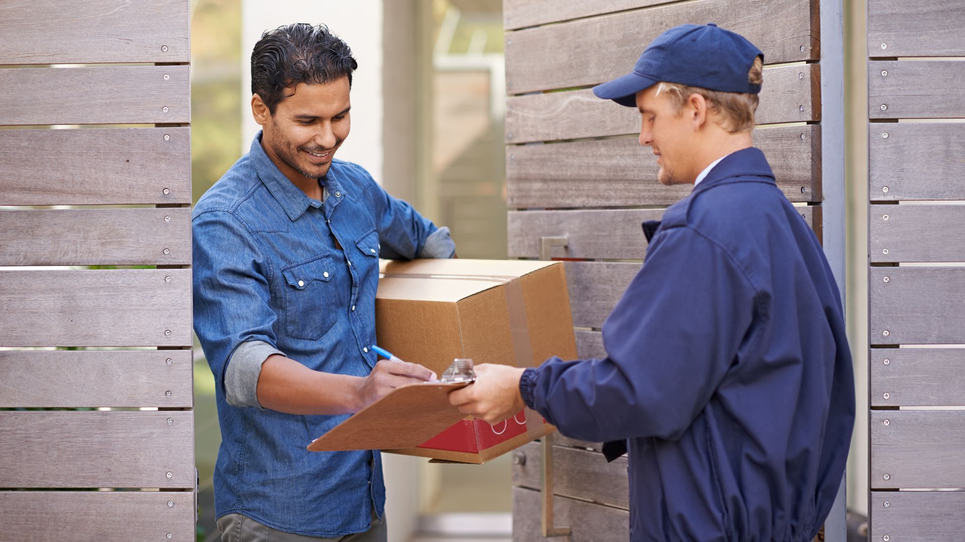 Delivery person hands a cardboard box to a person, both smiling, at a wooden doorway.