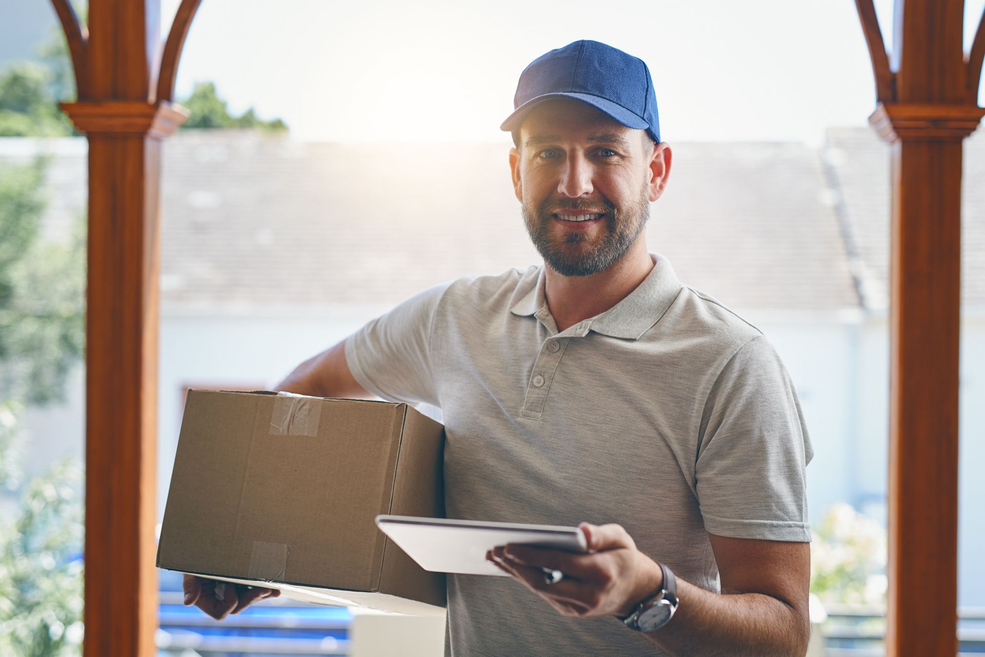 Delivery person holding a package and tablet, smiling at the camera outdoors.
