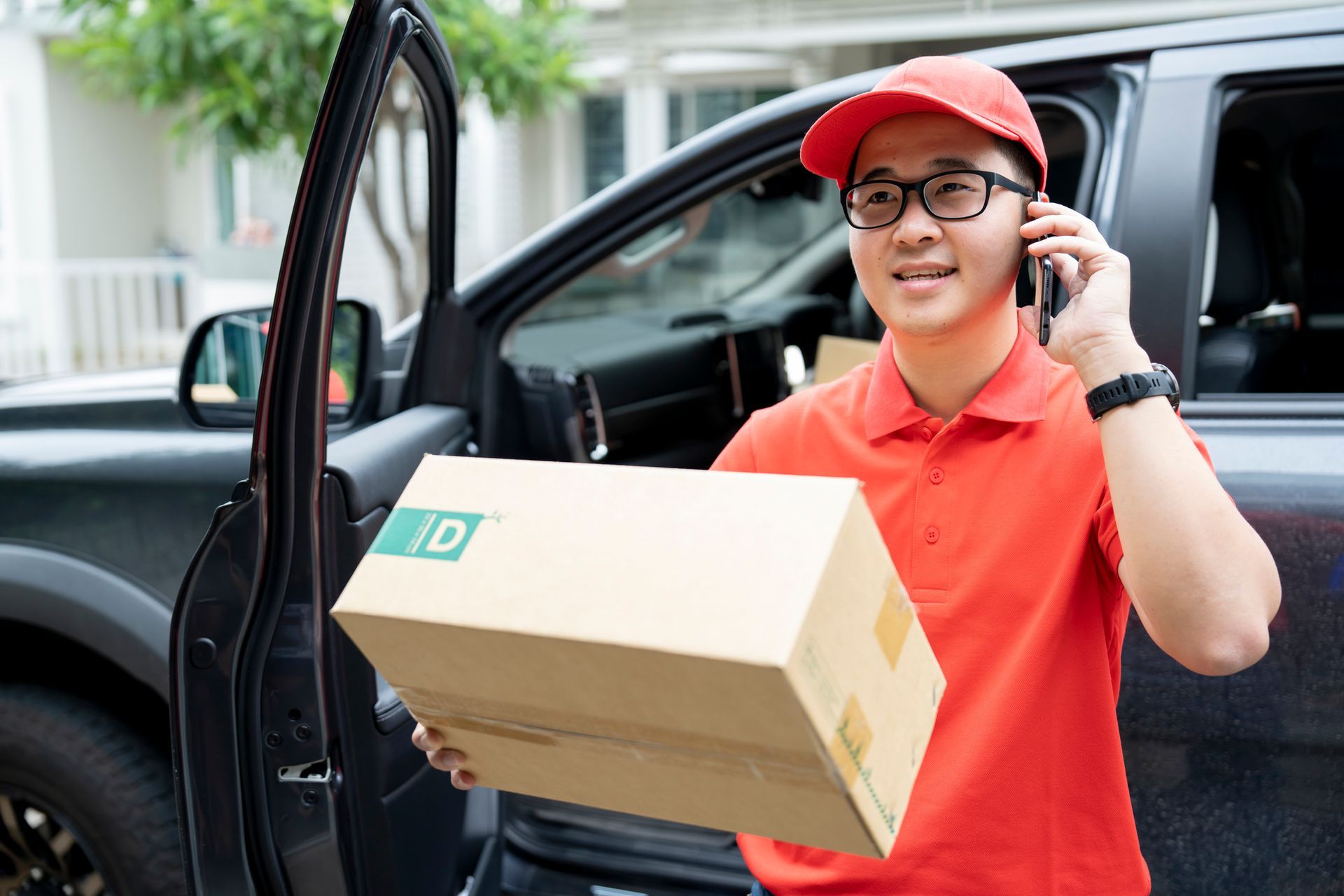 An Asian delivery man in a red uniform holding a cardboard package while talking on a mobile phone An Asian delivery man in a red uniform holding a cardboard package while talking on a mobile phone