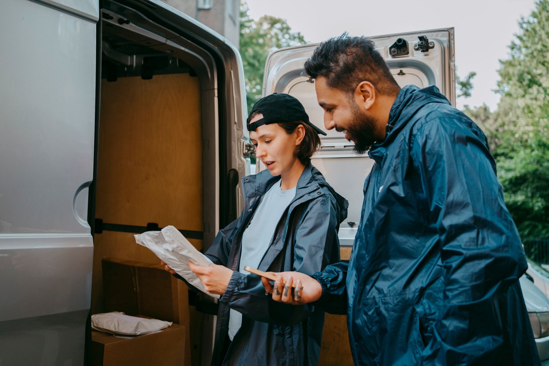Two people in rain jackets review papers next to a van full of boxes, outdoors. Two people in rain jackets review papers next to a van full of boxes, outdoors.