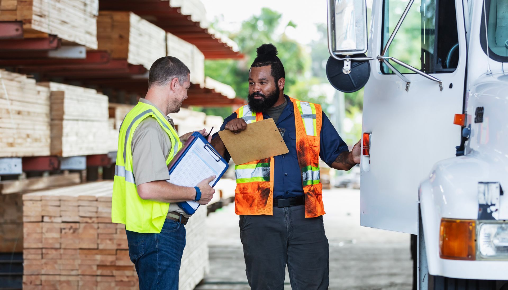 Two men in safety vests discussing paperwork next to a semi-truck in a lumberyard.