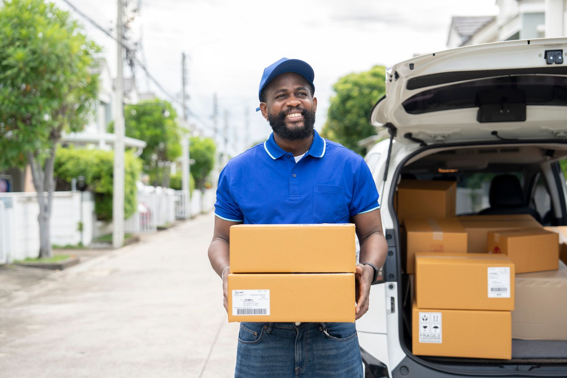 Delivery person holding packages near van, smiling. Blue shirt, cap. Suburban street. Delivery person holding packages near van, smiling. Blue shirt, cap. Suburban street.