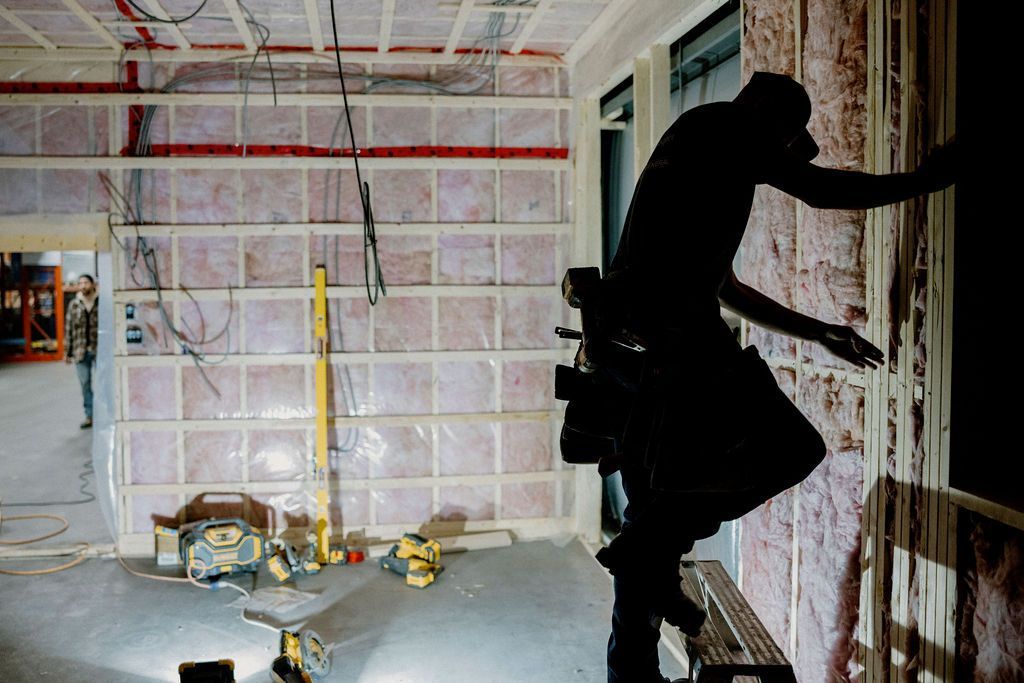 Silhouette d'un ouvrier du bâtiment travaillant sur une fenêtre, à l'intérieur. Isolation et outils visibles dans la pièce.