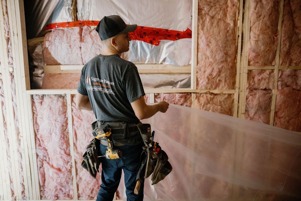 Un ouvrier du bâtiment installe une bâche en plastique sur un mur à ossature, avec une isolation rose visible.