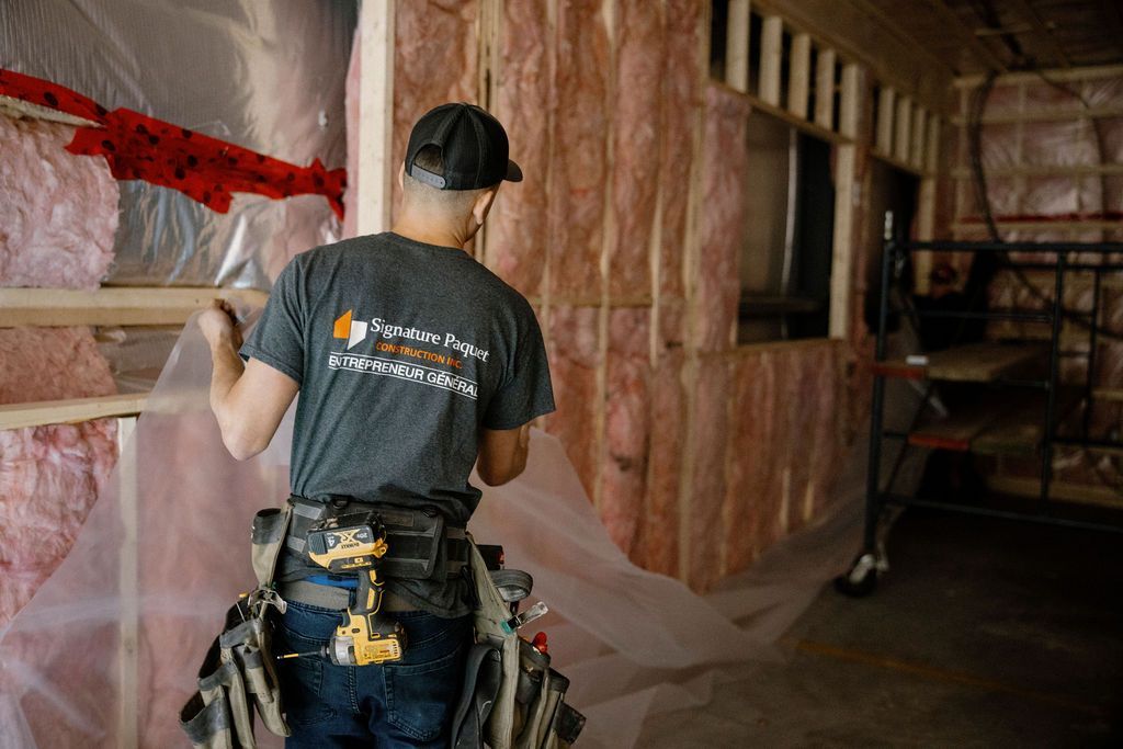 Un homme portant une ceinture à outils installe une bâche en plastique sur une isolation rose dans la charpente d'un bâtiment.