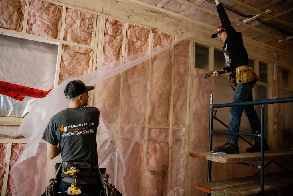 Deux ouvriers du bâtiment installent une bâche en plastique sur un mur avec une isolation rose. L'un d'eux est sur un échafaudage.