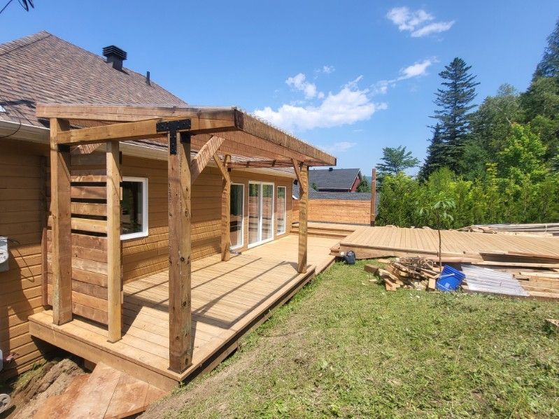 Terrasse en bois avec pergola attenante à une maison, journée ensoleillée, avec matériaux de construction à proximité.