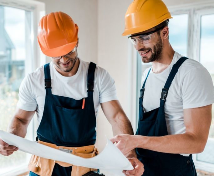 Deux ouvriers du bâtiment portant des casques et des combinaisons examinent un plan.