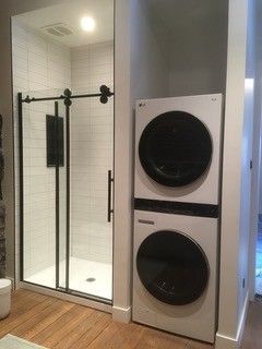 Salle de bain moderne : Douche avec cadre noir, à côté d'une laveuse et d'une sécheuse blanches superposées sur un plancher en bois.