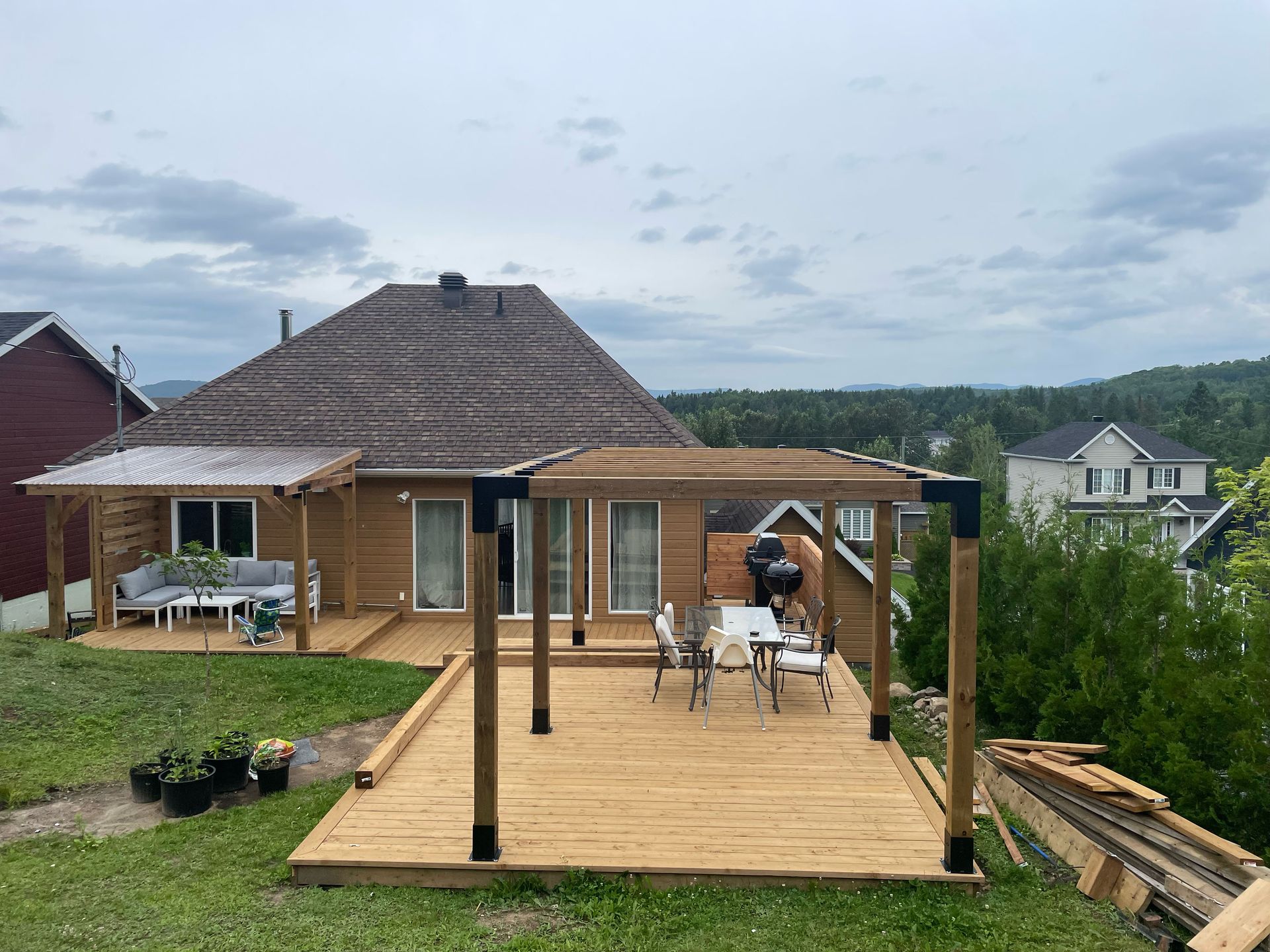Terrasse en bois avec pergola, ensemble de salle à manger et barbecue dans une cour arrière avec une maison et un paysage environnant.