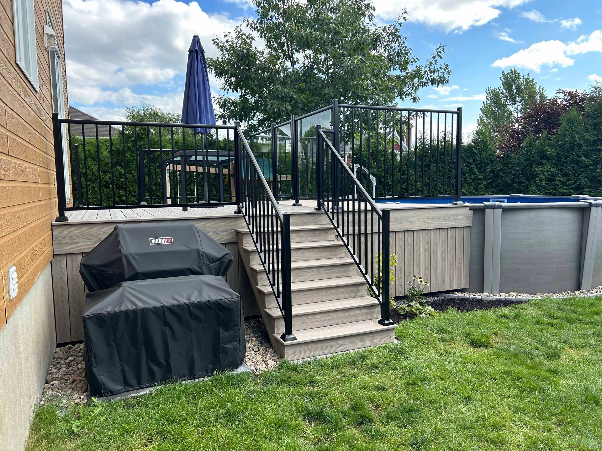 Terrasse arrière avec escalier, rampes noires et piscine hors sol. Pelouse verte et ciel bleu.