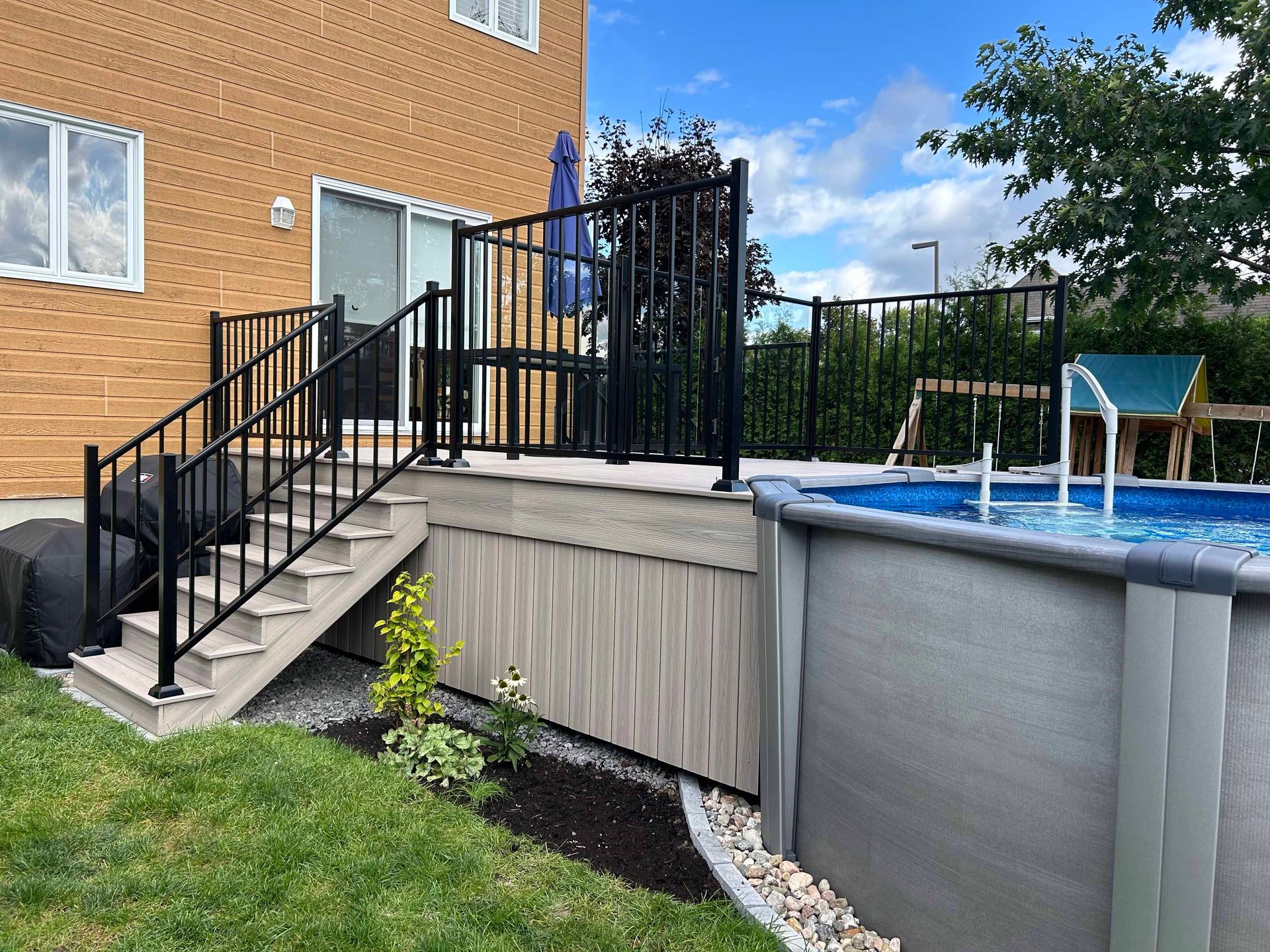 Terrasse arrière avec rampe noire, escalier et piscine hors sol. Herbe verte, ciel bleu et maison en briques.