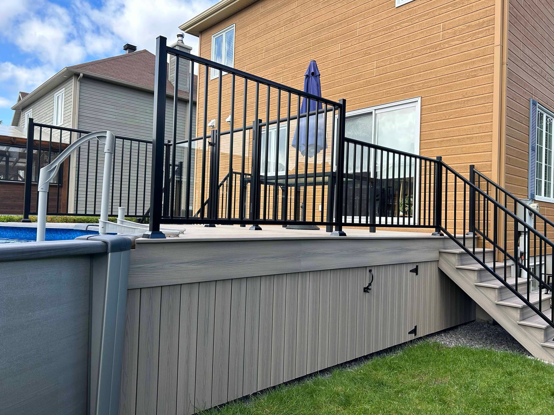 Une balustrade noire entoure une terrasse avec un escalier menant à une cour gazonnée. Une maison beige en arrière-plan.