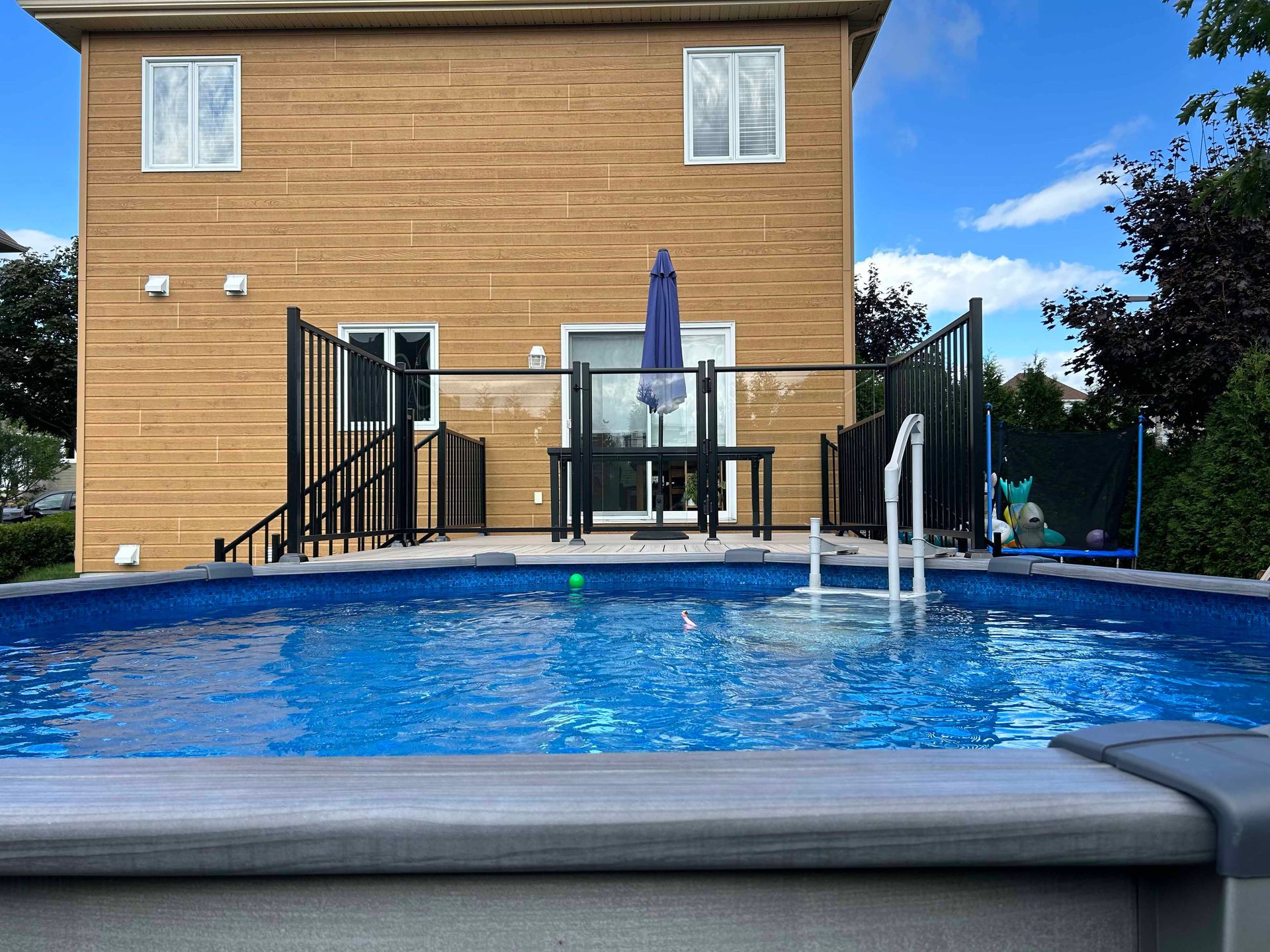 Jardin avec piscine hors sol. Une maison au revêtement brun clair se dresse à l'arrière-plan. Ciel bleu.