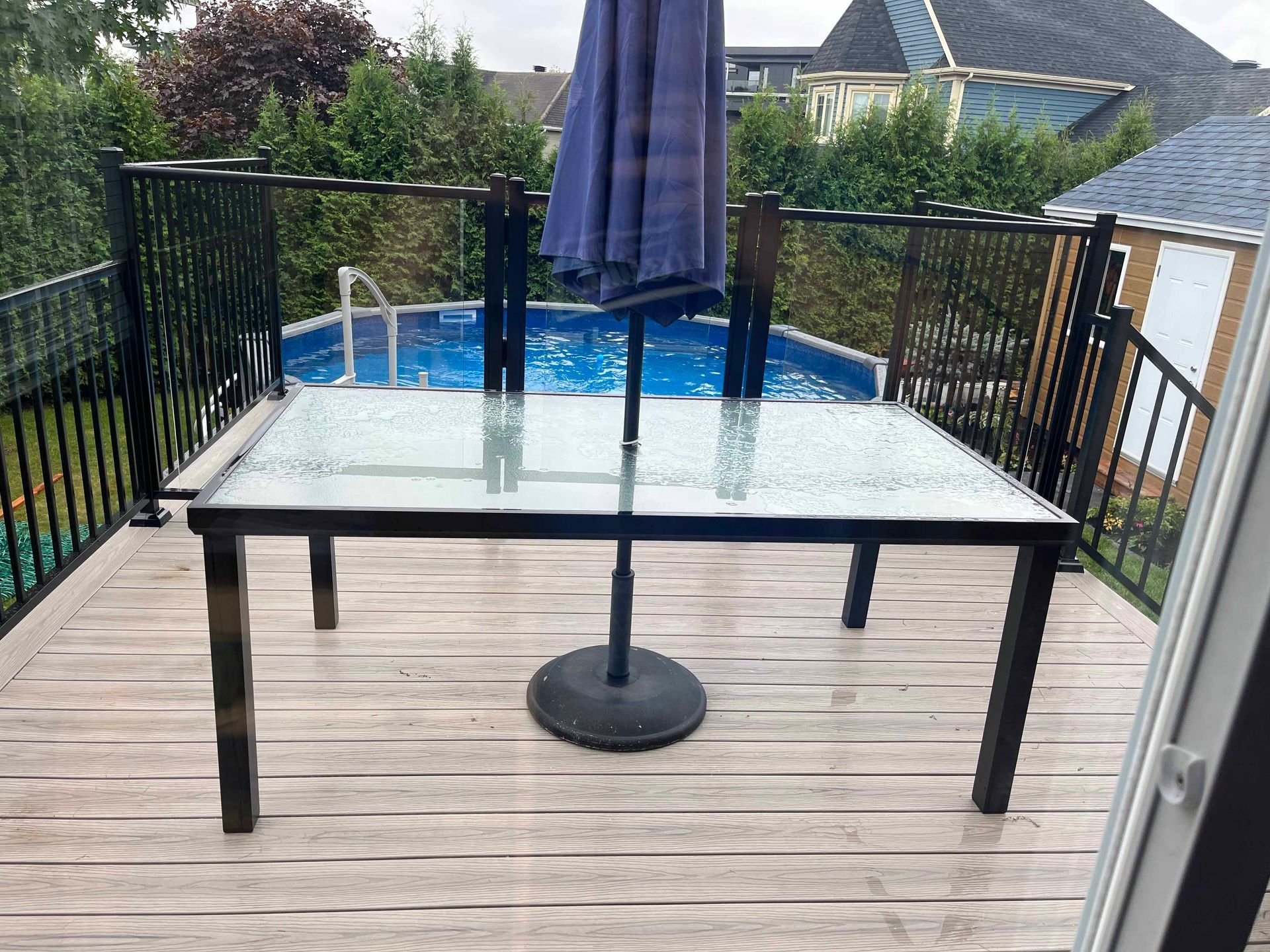 Terrasse extérieure avec une table en verre, un parasol bleu et une petite piscine derrière une balustrade noire.