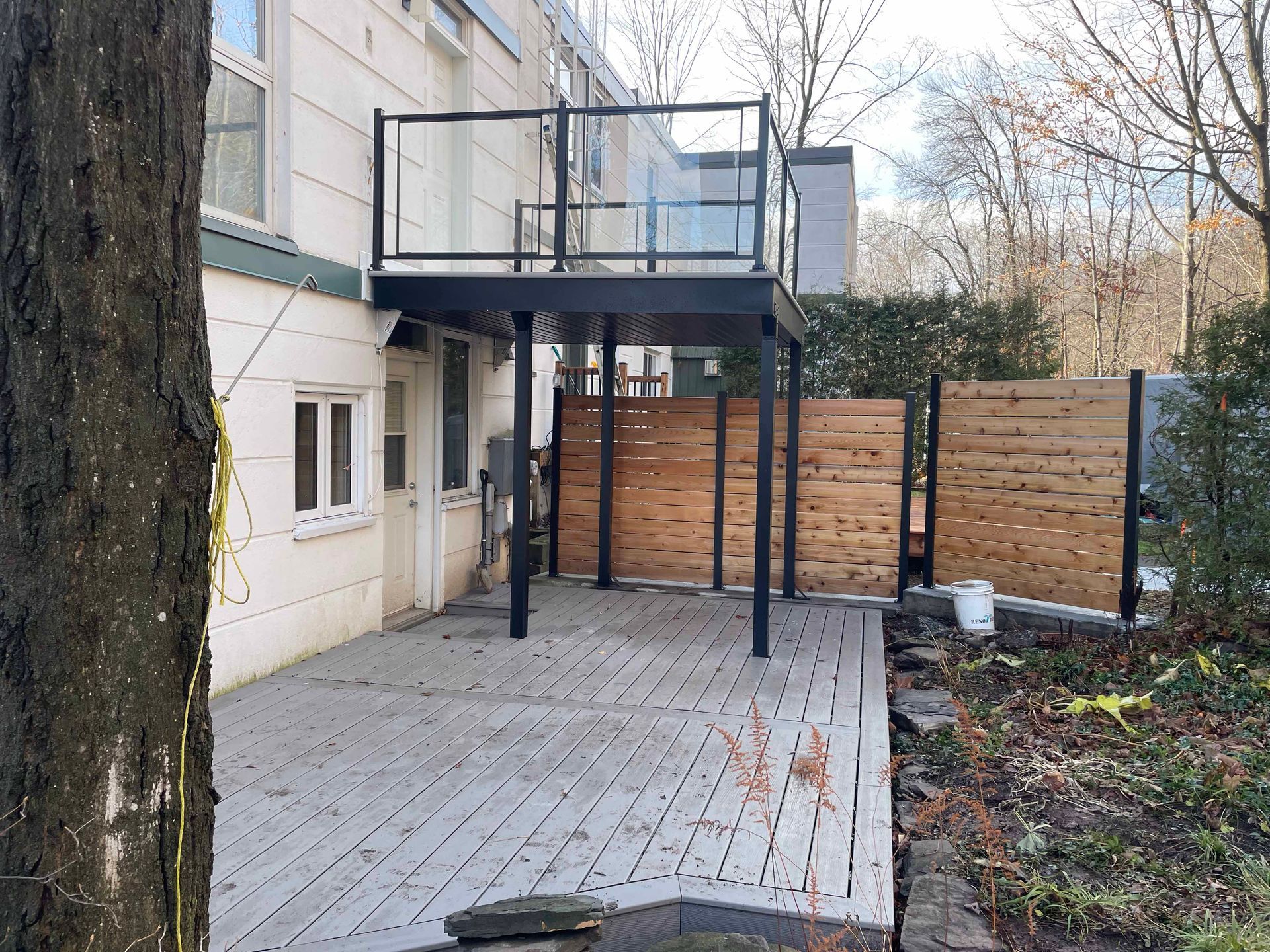 Terrasse en bois gris avec un balcon noir et en verre, une clôture d'intimité marron et un arbre.
