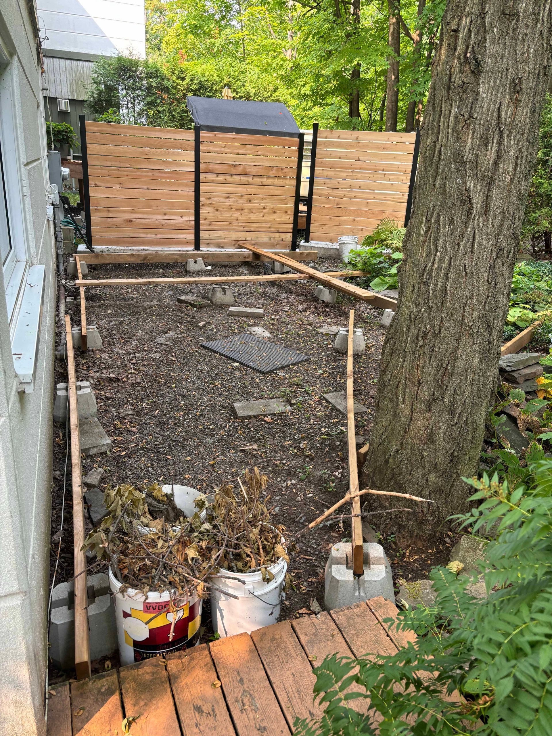 Clôture en bois, chemin et arbre dans un espace extérieur. Seaux de plantes séchées et pierres de gué.