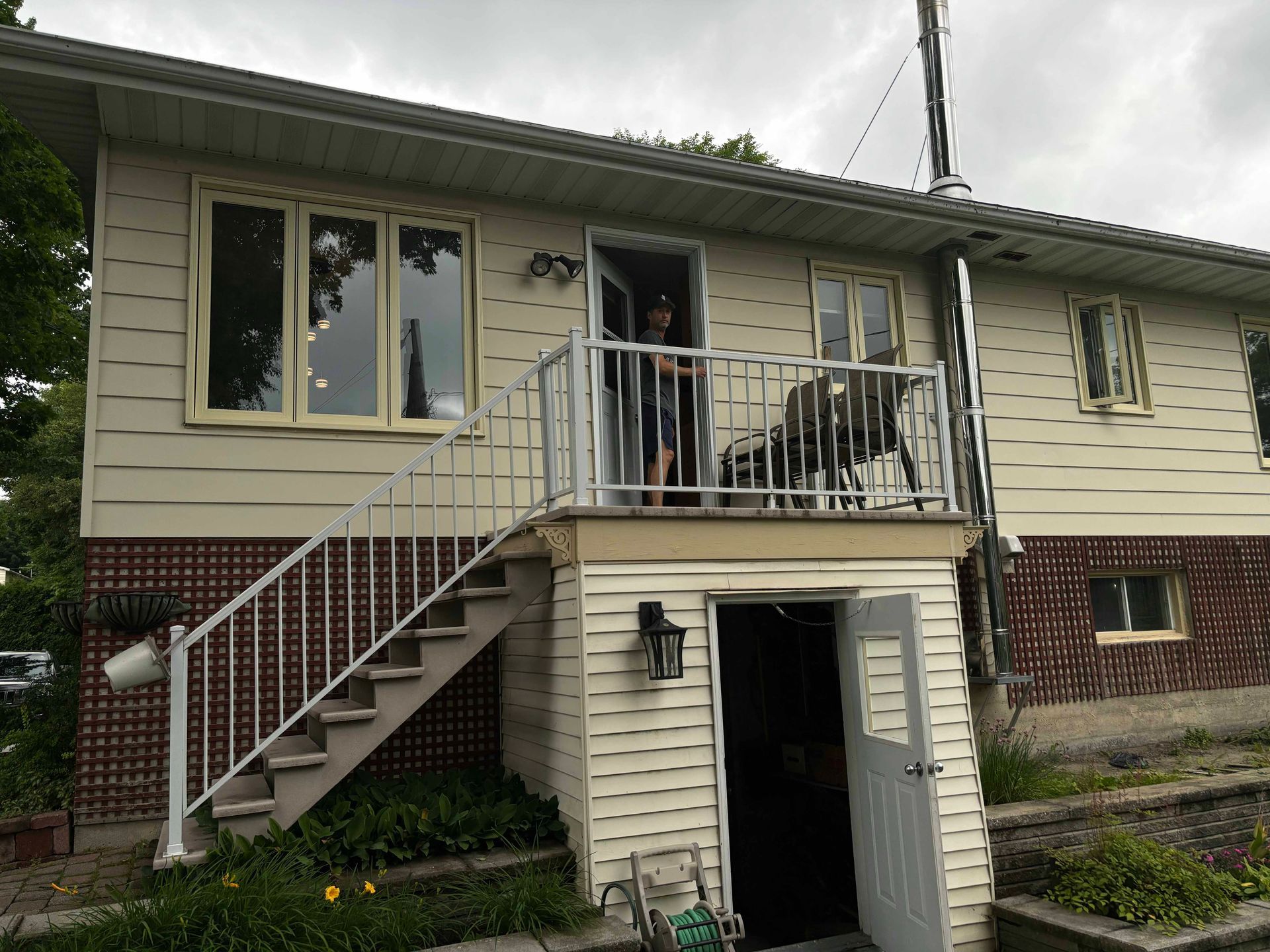 Maison à deux étages avec escalier extérieur menant à l'étage. Une personne se tient dans l'embrasure d'une porte sur la terrasse.