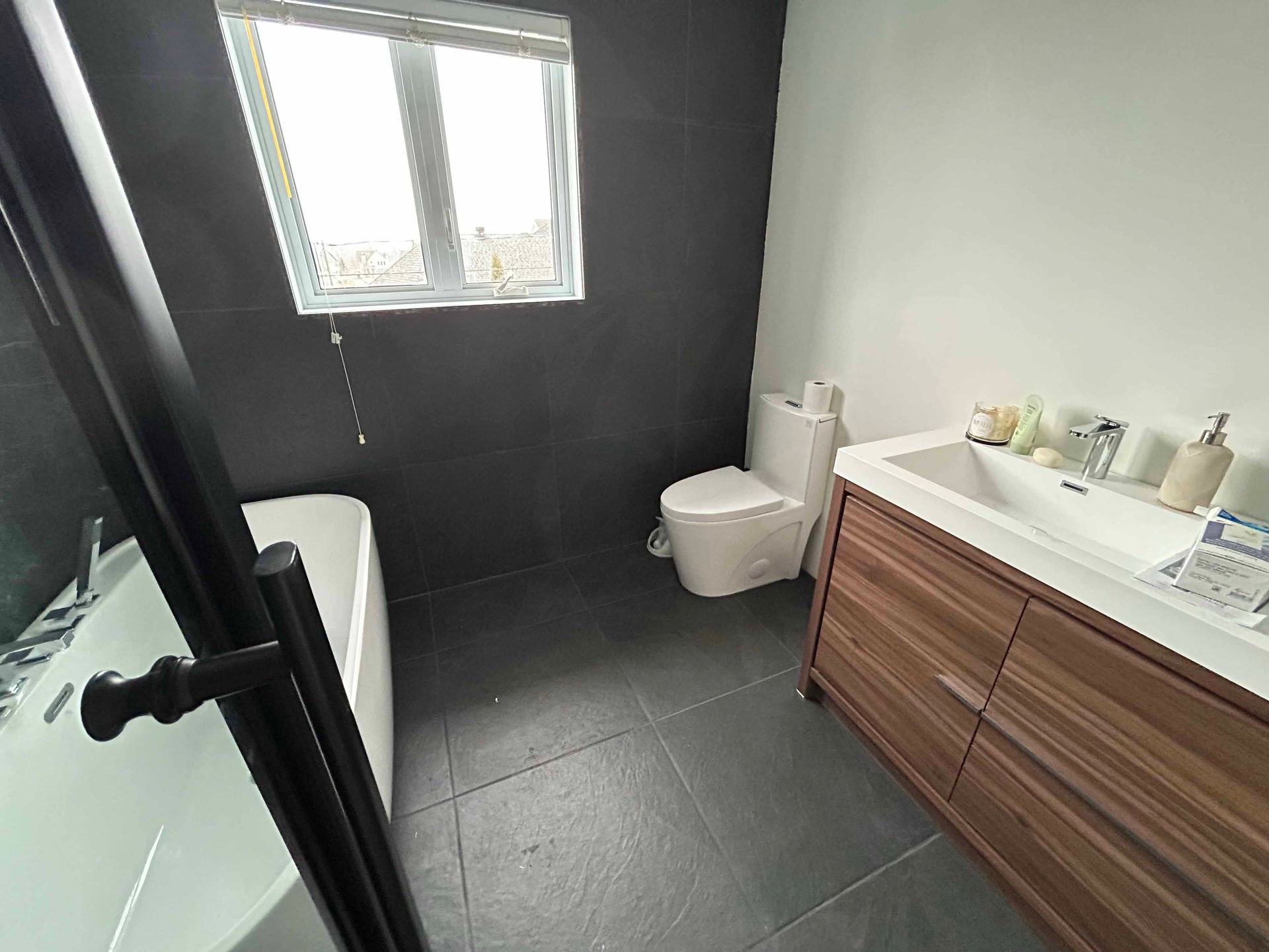 Salle de bain avec carrelage foncé, luminaires blancs, vanité en bois et fenêtre.