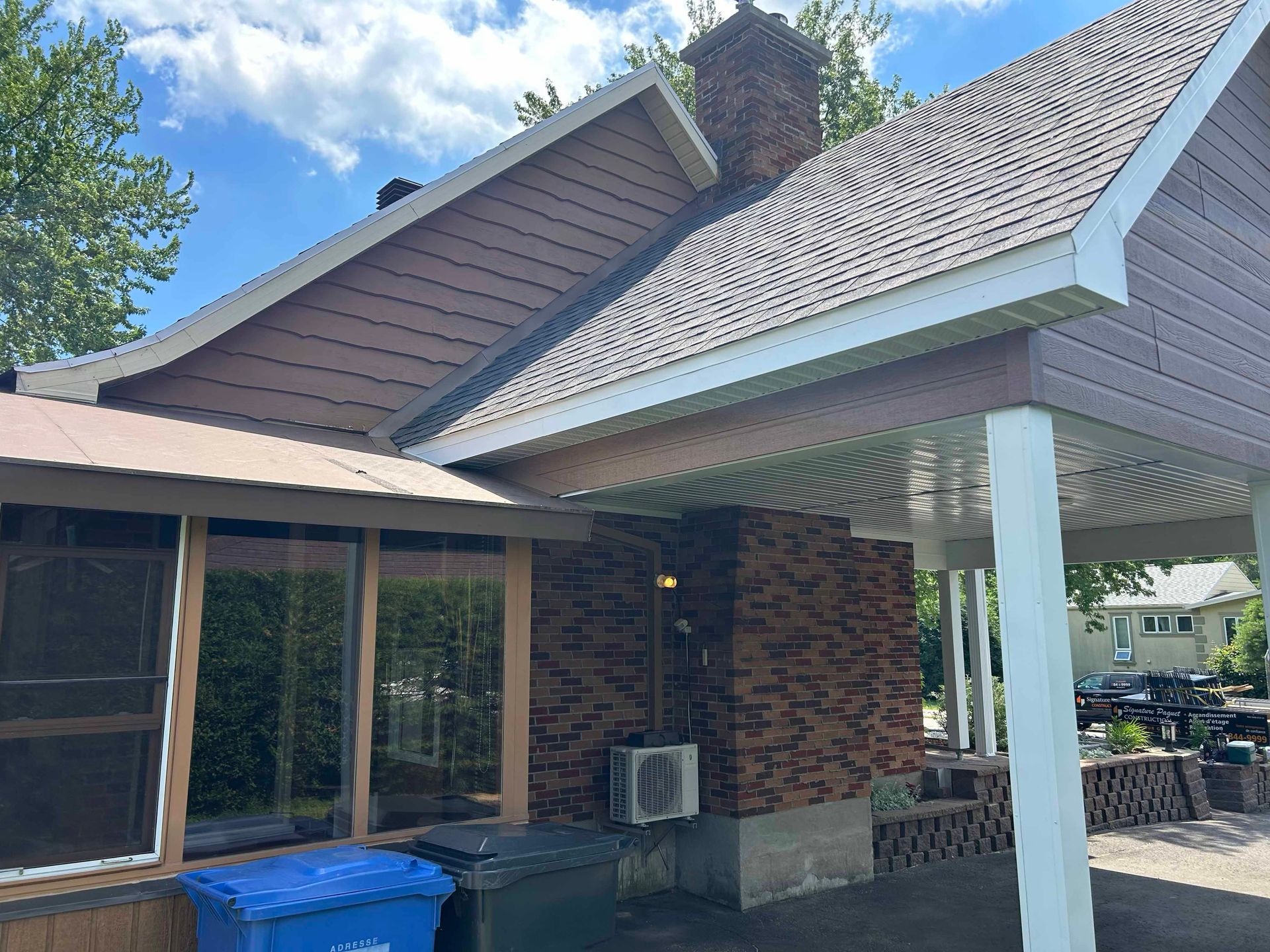 Extérieur de maison en brique avec revêtement marron, porche couvert et bac de recyclage bleu.