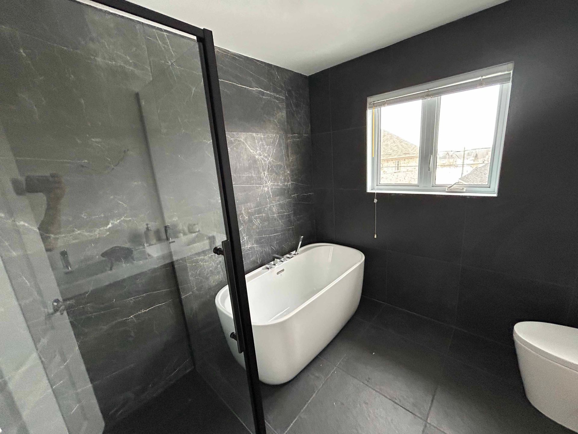 Salle de bain moderne avec murs et sol en carrelage noir, baignoire blanche et douche en verre.