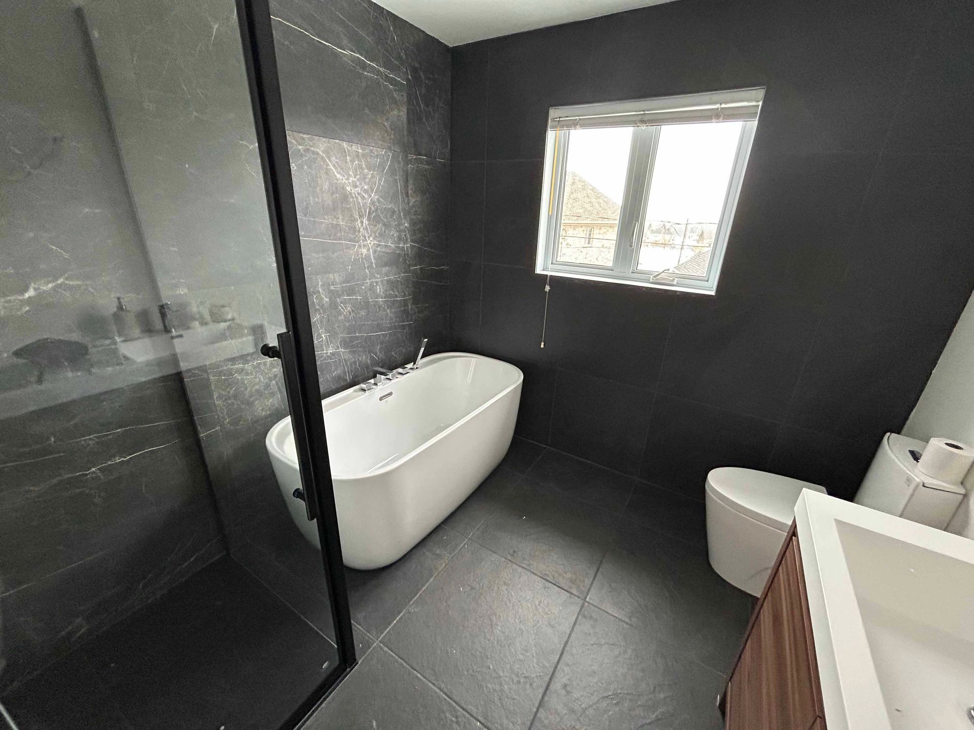 Salle de bain moderne avec carrelage gris foncé aux murs et au sol. Baignoire îlot, toilettes et lavabo blancs.