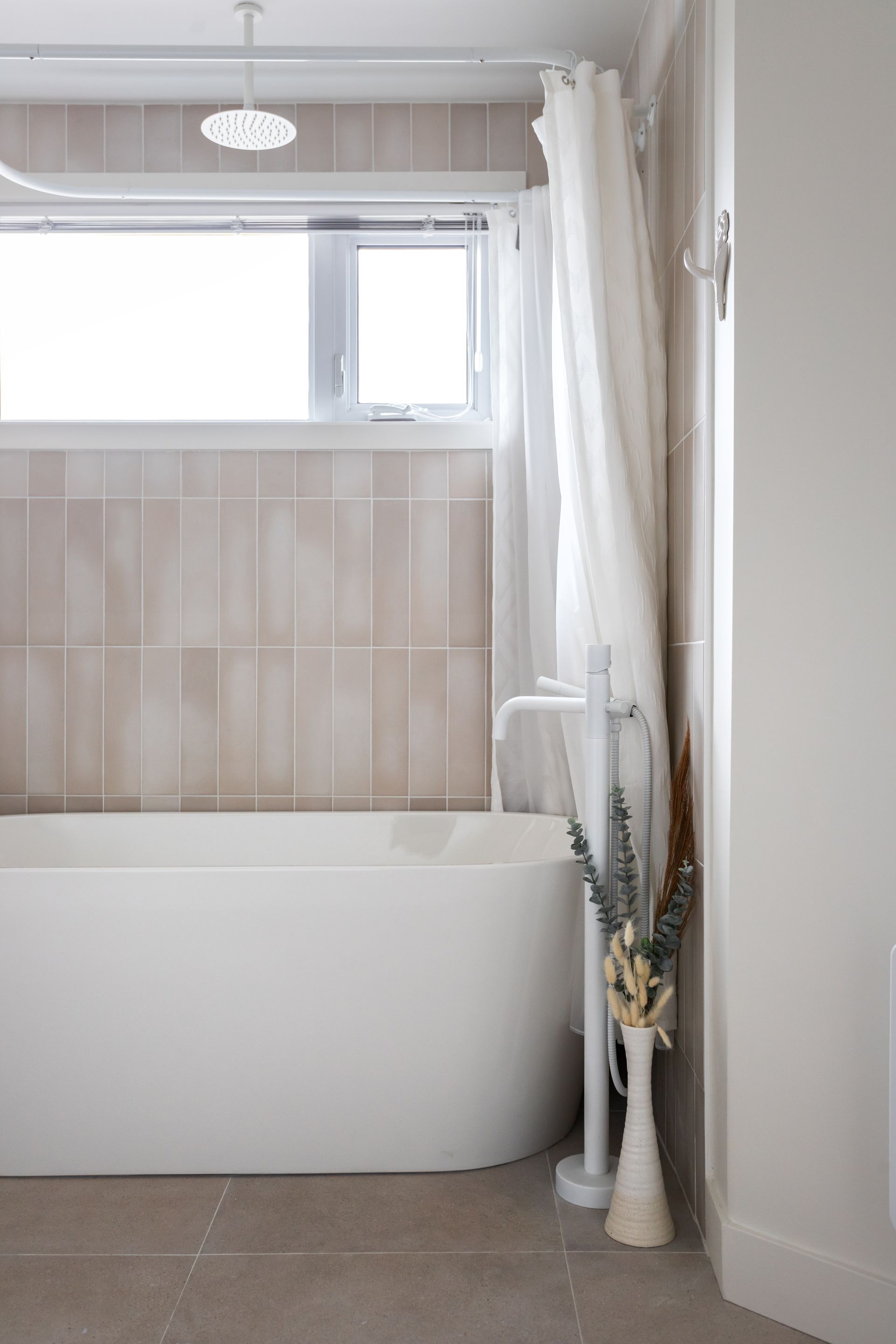 Baignoire blanche sous une fenêtre dans une salle de bain aux tons neutres. Un rideau blanc et un vase de fleurs séchées se trouvent à côté.