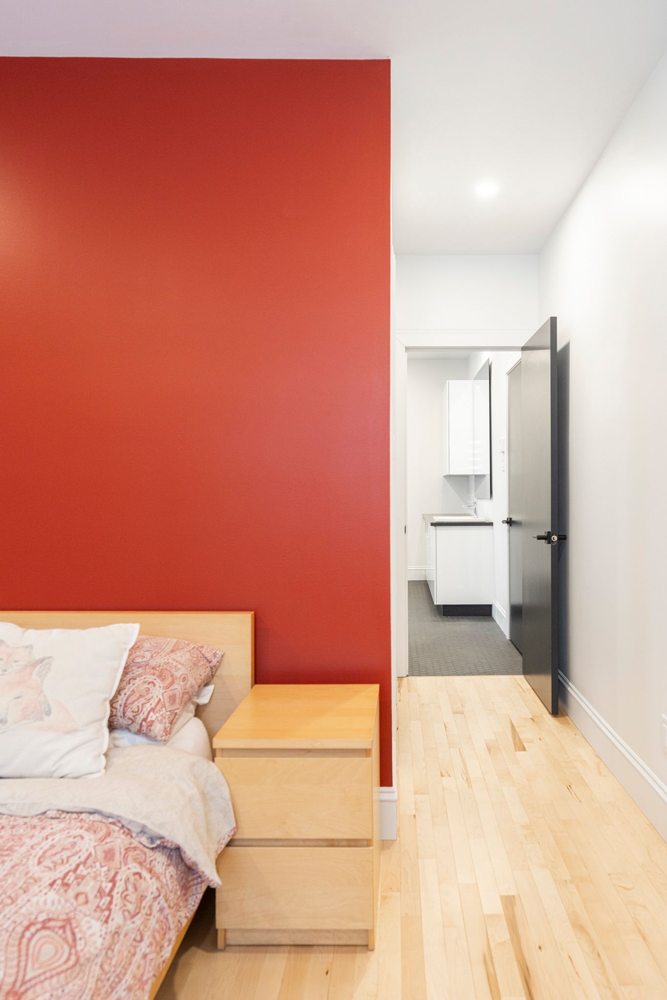 Chambre avec un mur d'accent rouge, une table de chevet en bois et une porte ouverte sur un couloir aux murs blancs.