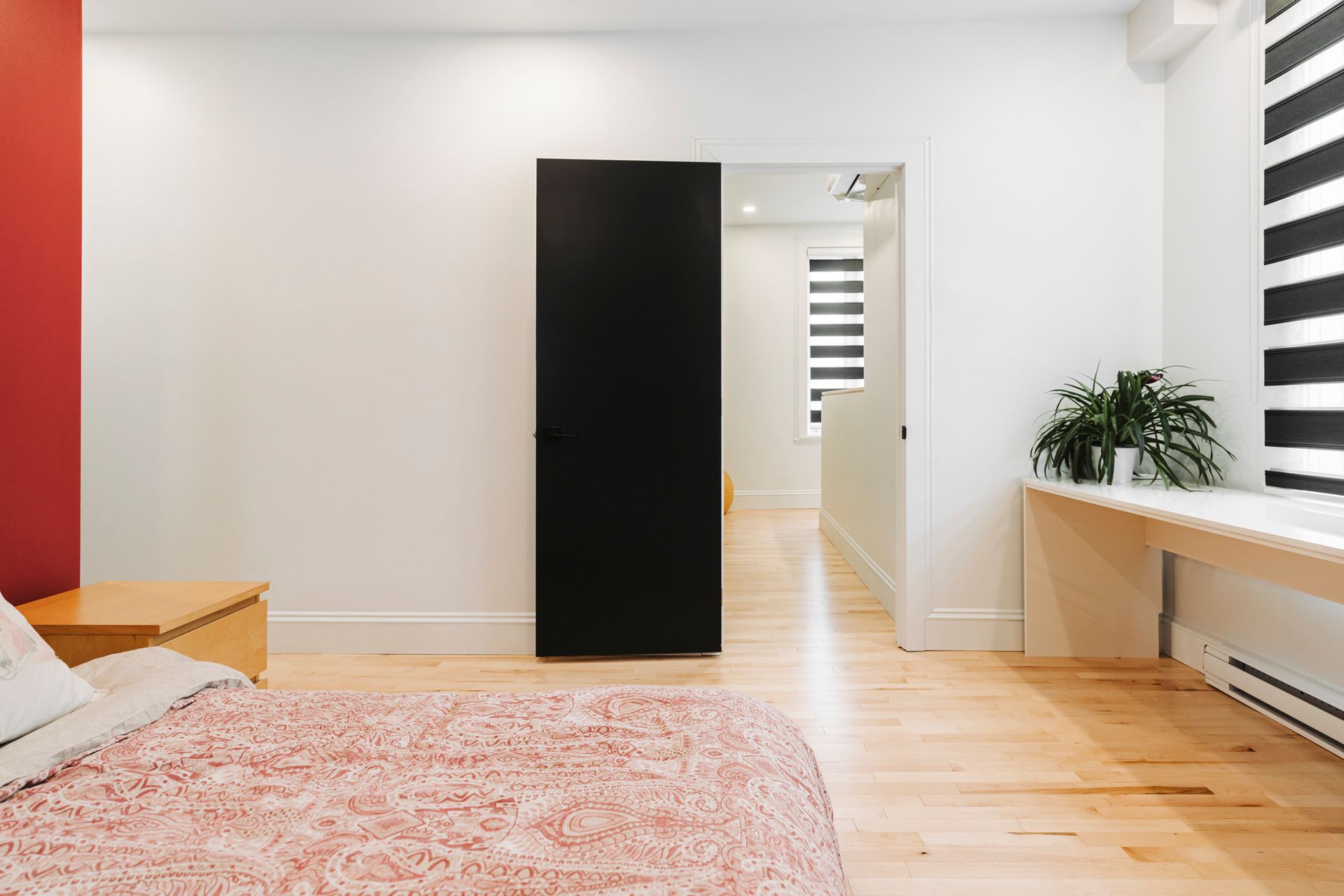 Chambre avec un mur d'accent rouge, une porte noire, du parquet et un long banc blanc avec une plante.