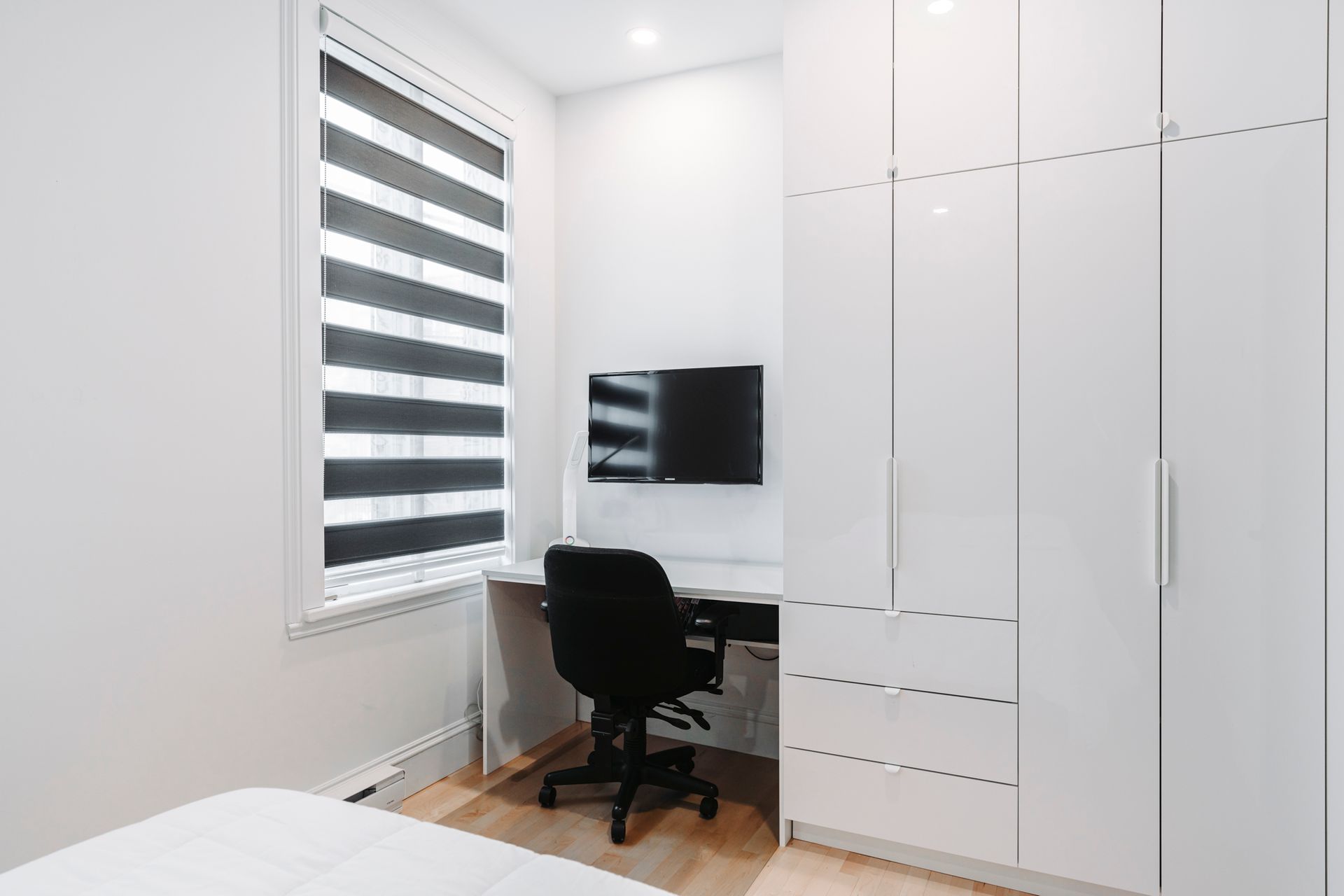 Chambre blanche avec bureau, chaise noire, armoire, fenêtre avec stores zébrés.