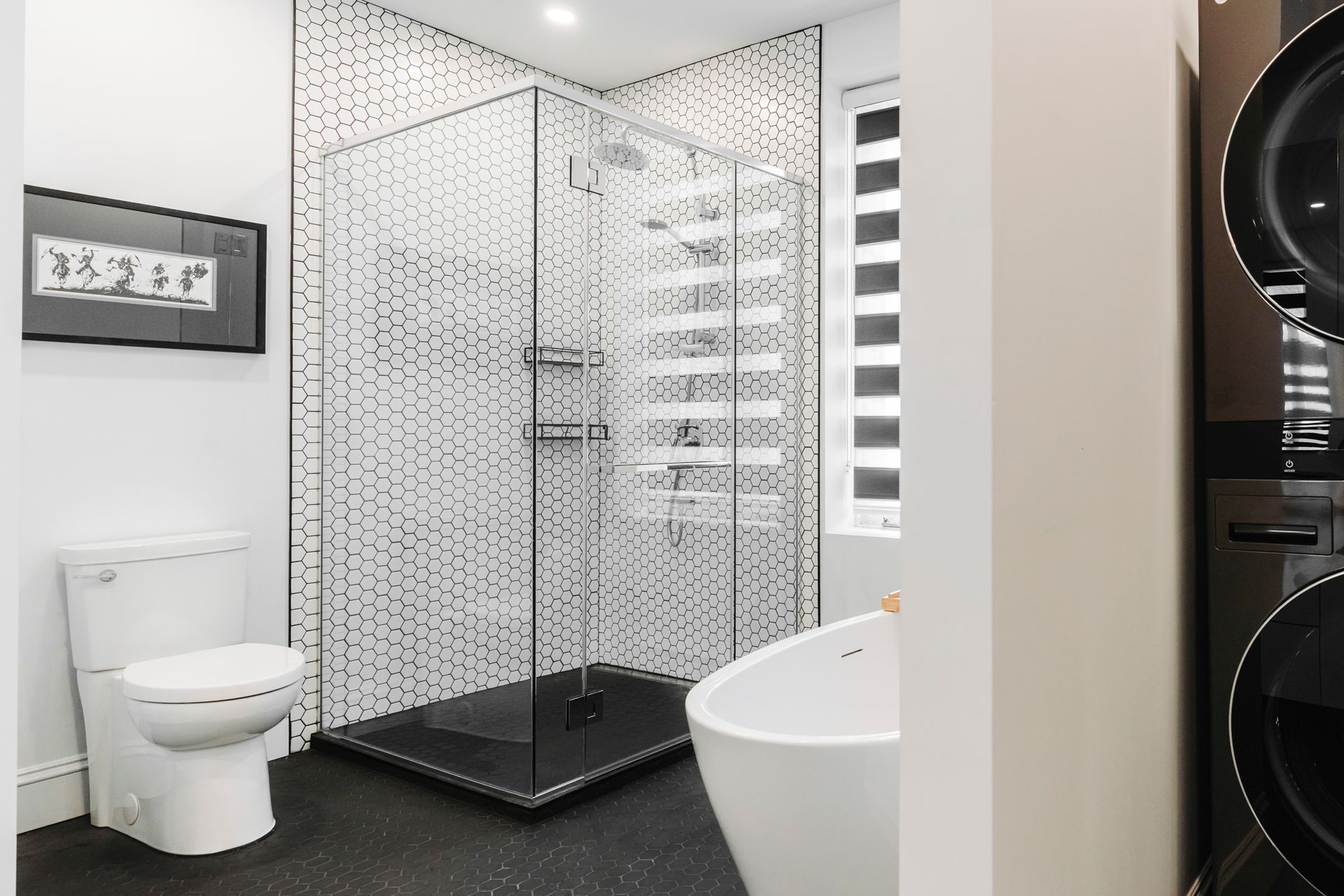 Salle de bain moderne avec douche carrelée, toilettes, baignoire et lave-linge/sèche-linge superposés. Couleurs noir, blanc et gris.