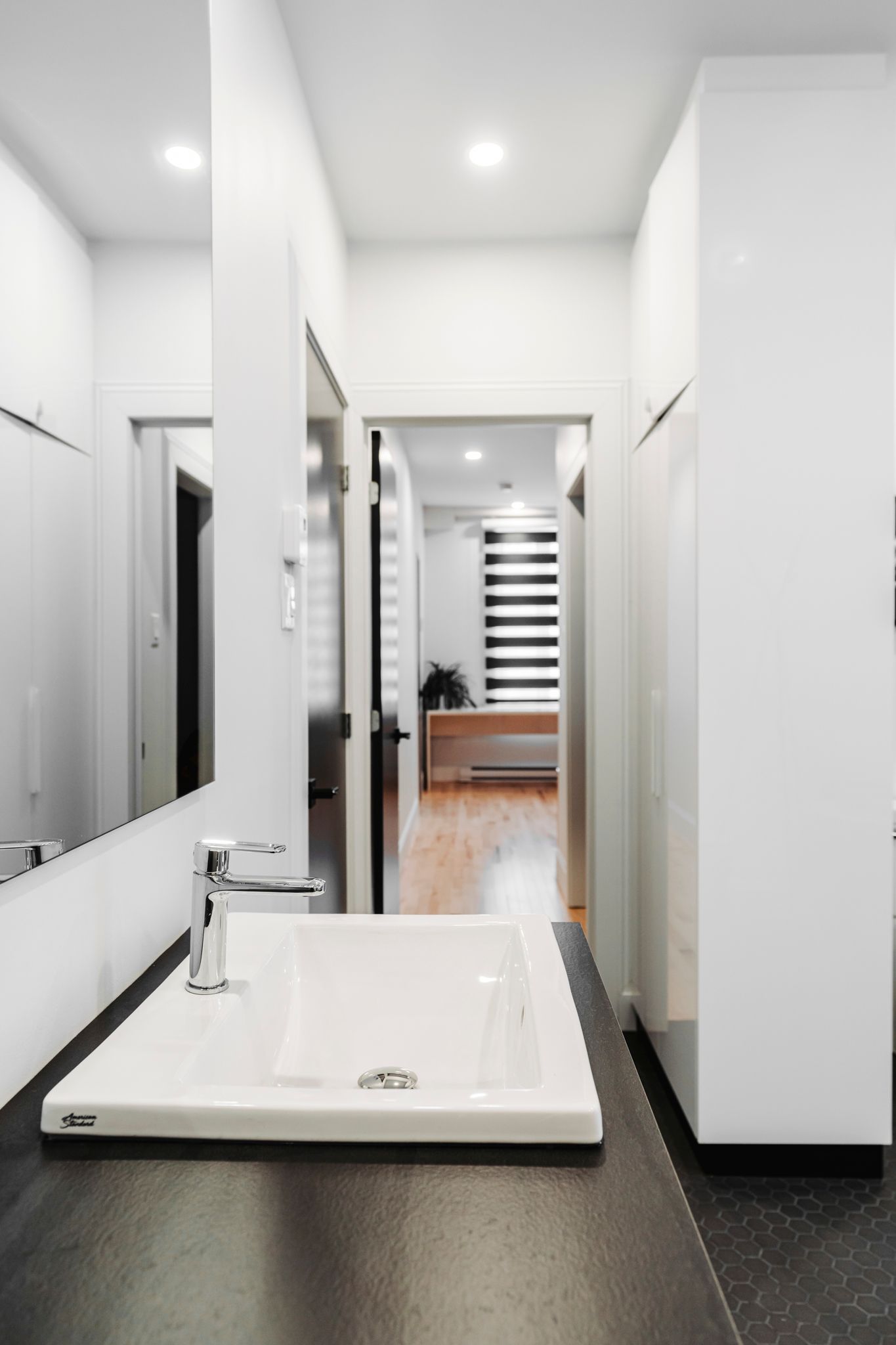 Salle de bain avec lavabo blanc, miroir et porte donnant sur un couloir avec une fenêtre.