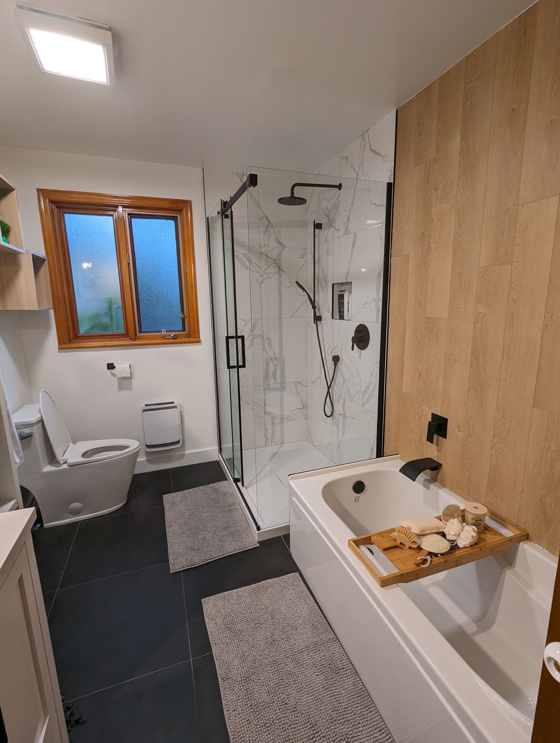 Salle de bain moderne avec sol en carrelage noir, baignoire blanche, douche en verre et accents en bois.