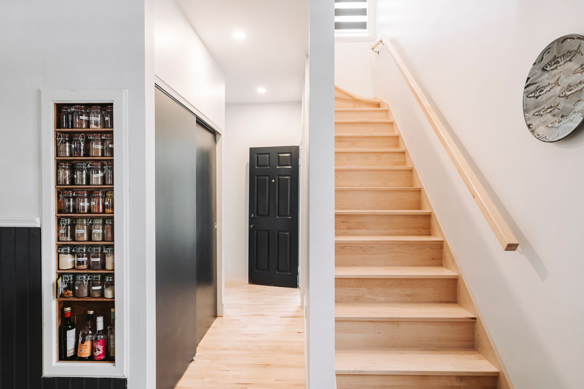 Couloir avec escalier en bois, étagère à épices, porte noire et murs et sol de couleur claire.