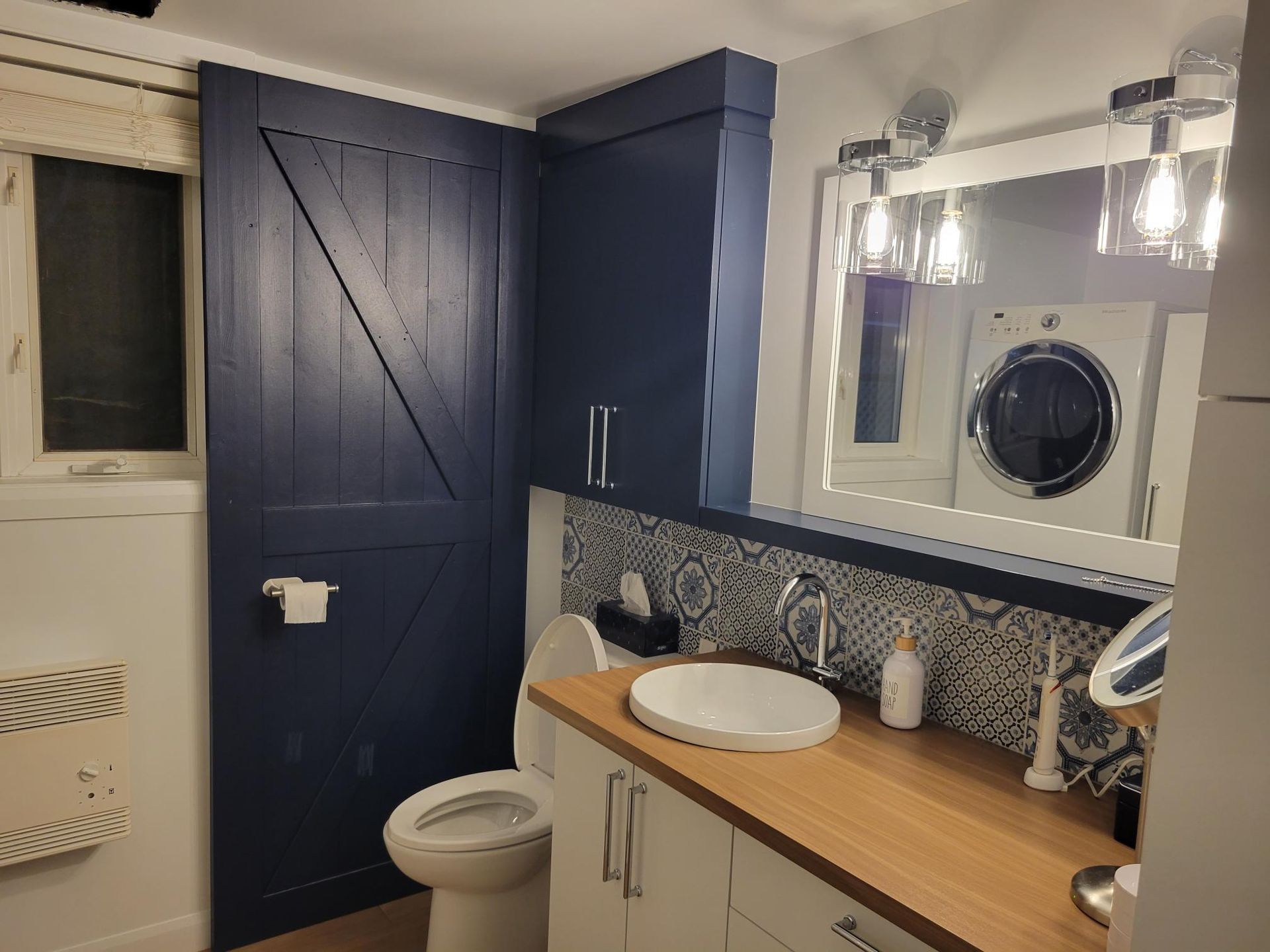 Salle de bain avec porte de grange bleu marine, vanité blanche, comptoir en bois et machine à laver dans le miroir.