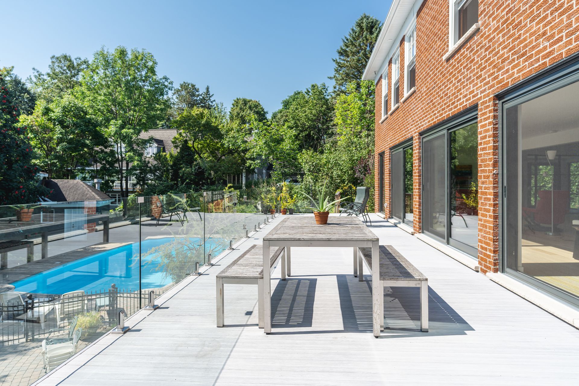 Extérieur d'une maison en briques avec piscine, terrasse en bois et table. Journée ensoleillée, arbres verdoyants.