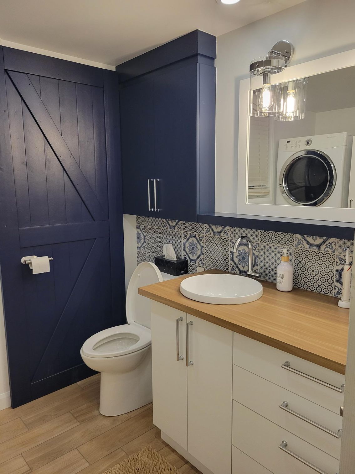 Une petite salle de bain moderne avec une porte de grange bleue, des armoires blanches et une machine à laver visible dans le miroir.
